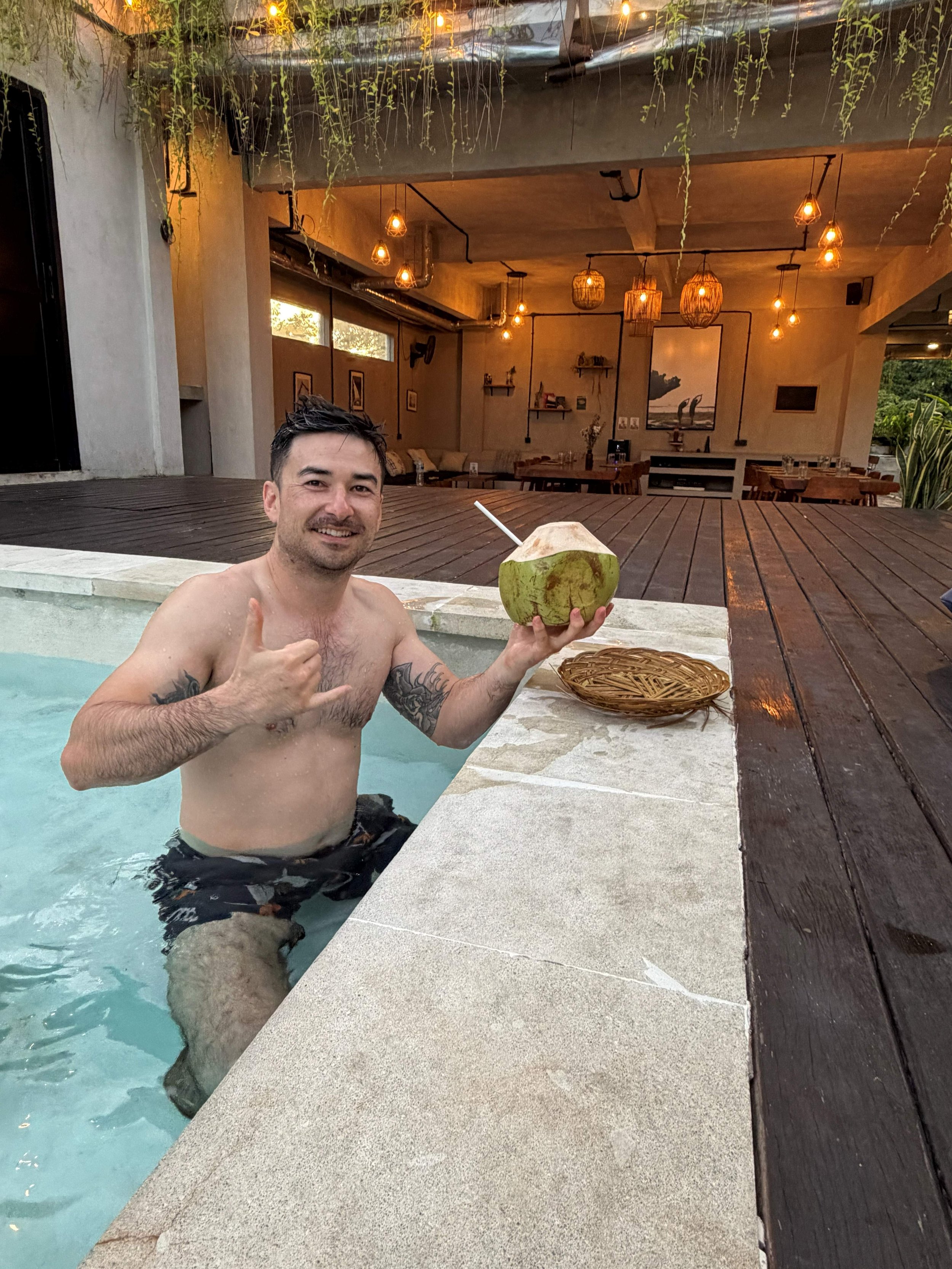 A man in a swimming pool holding a green coconut with a straw, giving a thumbs-up, with warm ambient lighting and a cozy indoor lounge area in the background.
