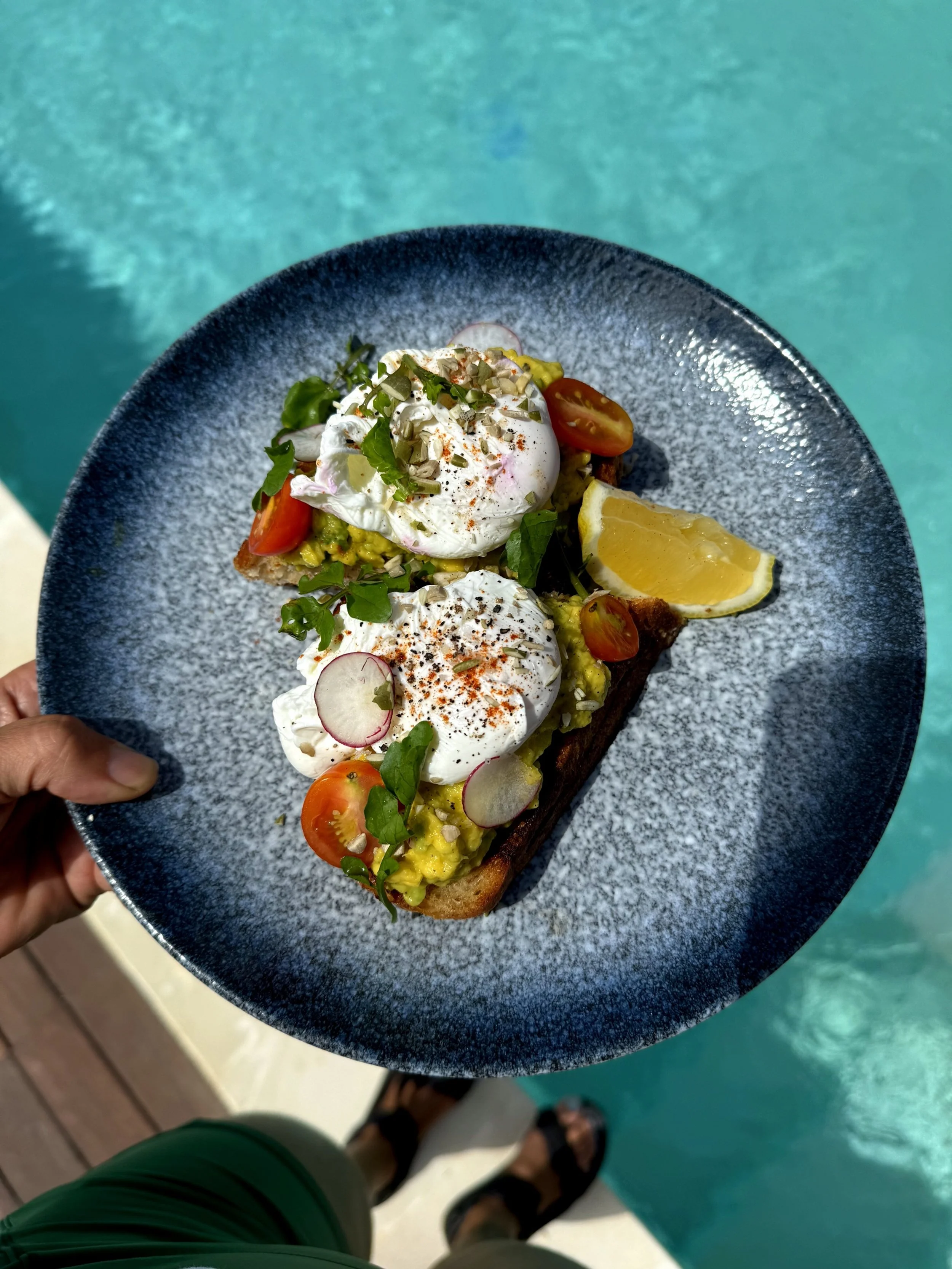 A person holding a plate with avocado toast topped with poached eggs, cherry tomatoes, radish slices, chopped nuts, and herbs, with a lemon wedge on the side, by a swimming pool.