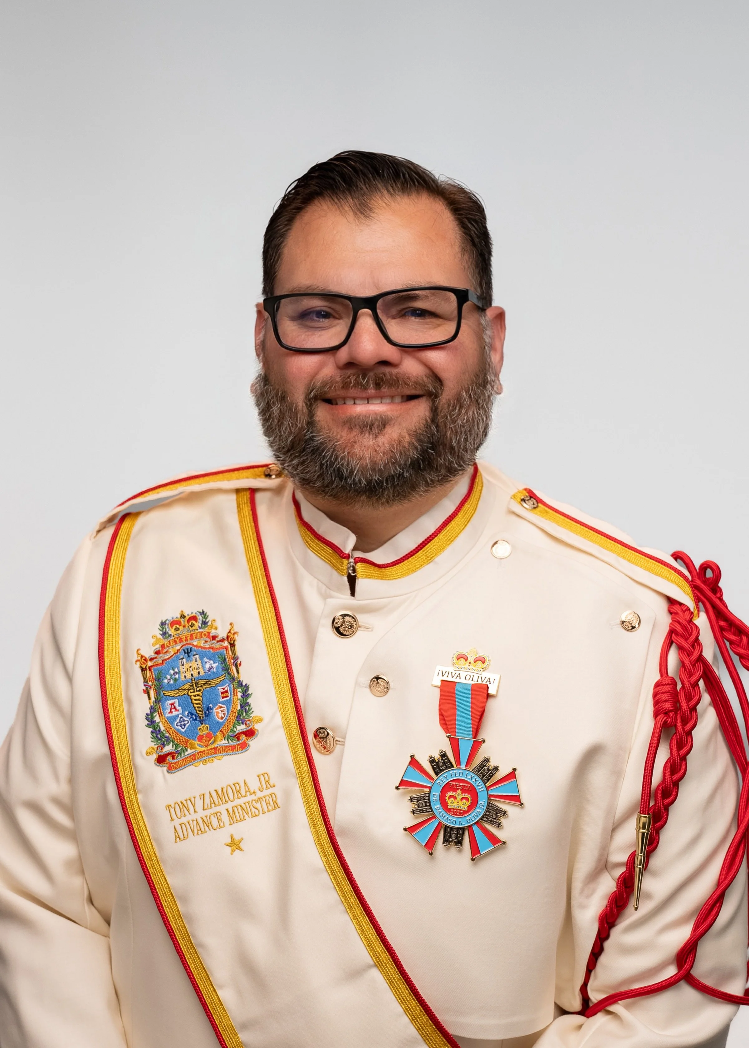 Close-up portrait of a smiling man with glasses, a beard, and dark hair. He is wearing a red button-up shirt and standing against a plain gray background.