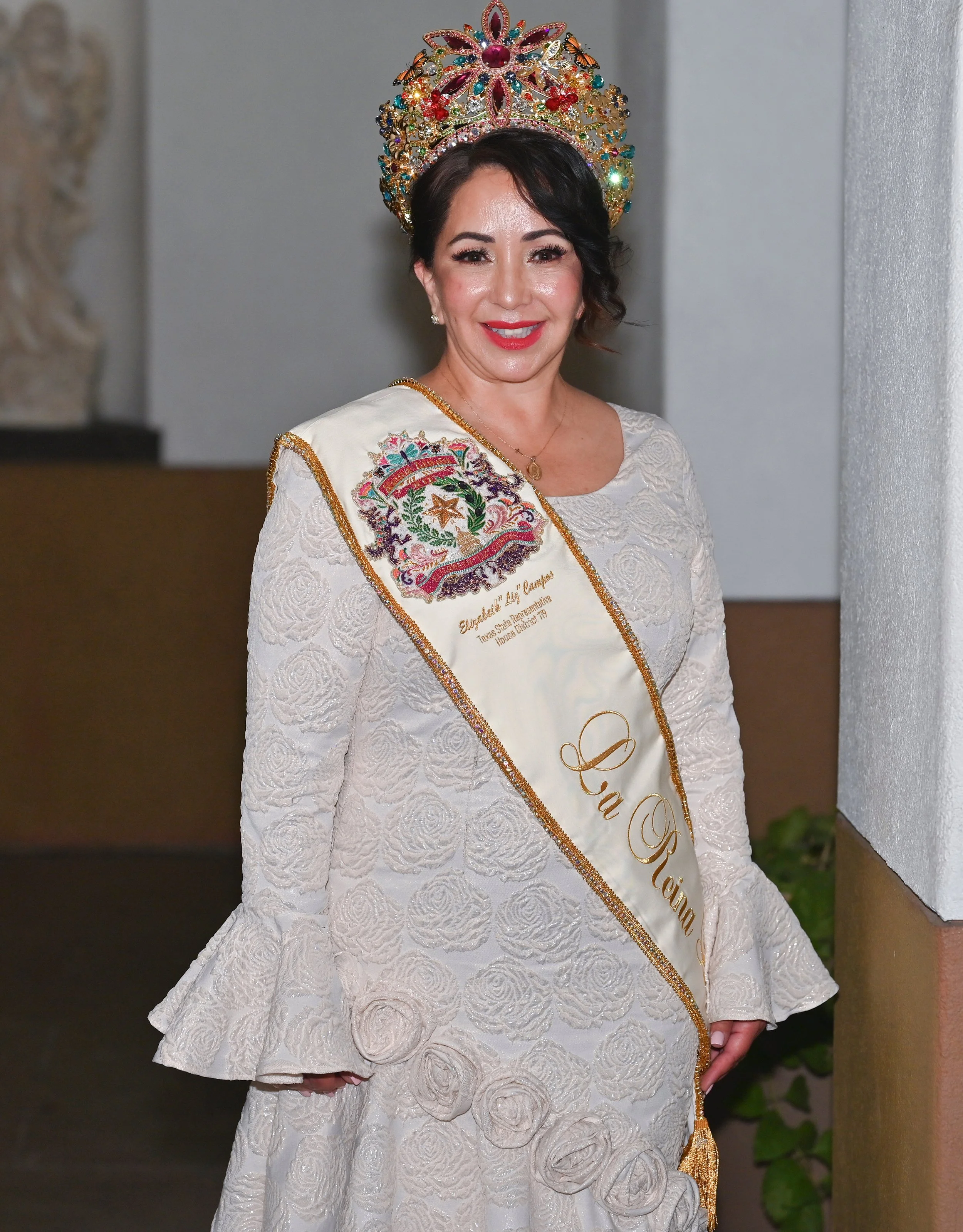 A woman with long, dark, wavy hair, wearing a sparkly, pink sequin top, smiling at the camera against a light gray background.