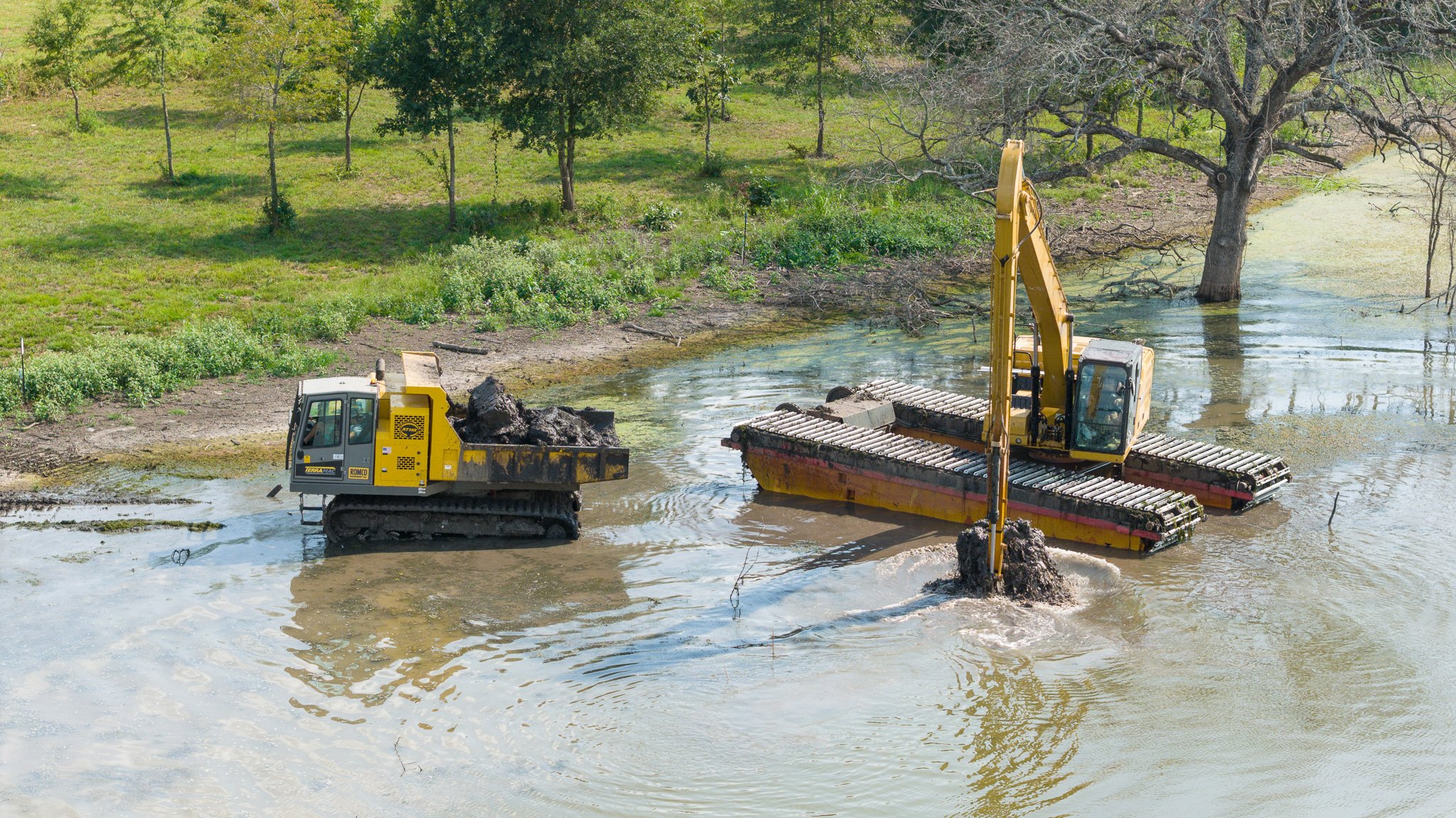 Construction machinery in a flooded area, with an excavator and a floating platform removing dirt and debris from the water near a tree and greenery.
