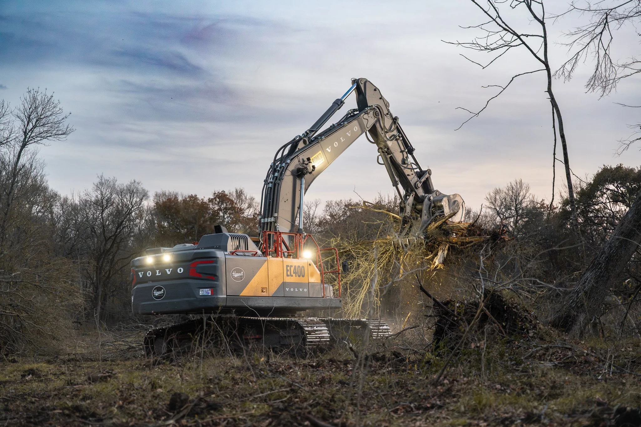 A Volvo excavator clearing trees and debris in a wooded area during dusk or early evening.