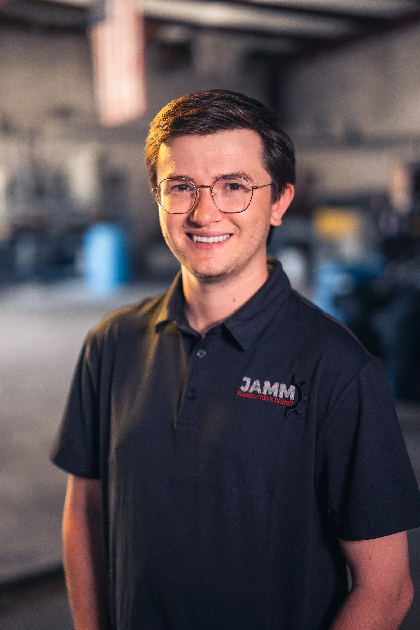 A young man with short dark hair, glasses, and a friendly smile, wearing a black polo shirt with 'JAMM' and 'Fabrication & Design' embroidered on it, standing in an industrial or workshop setting.