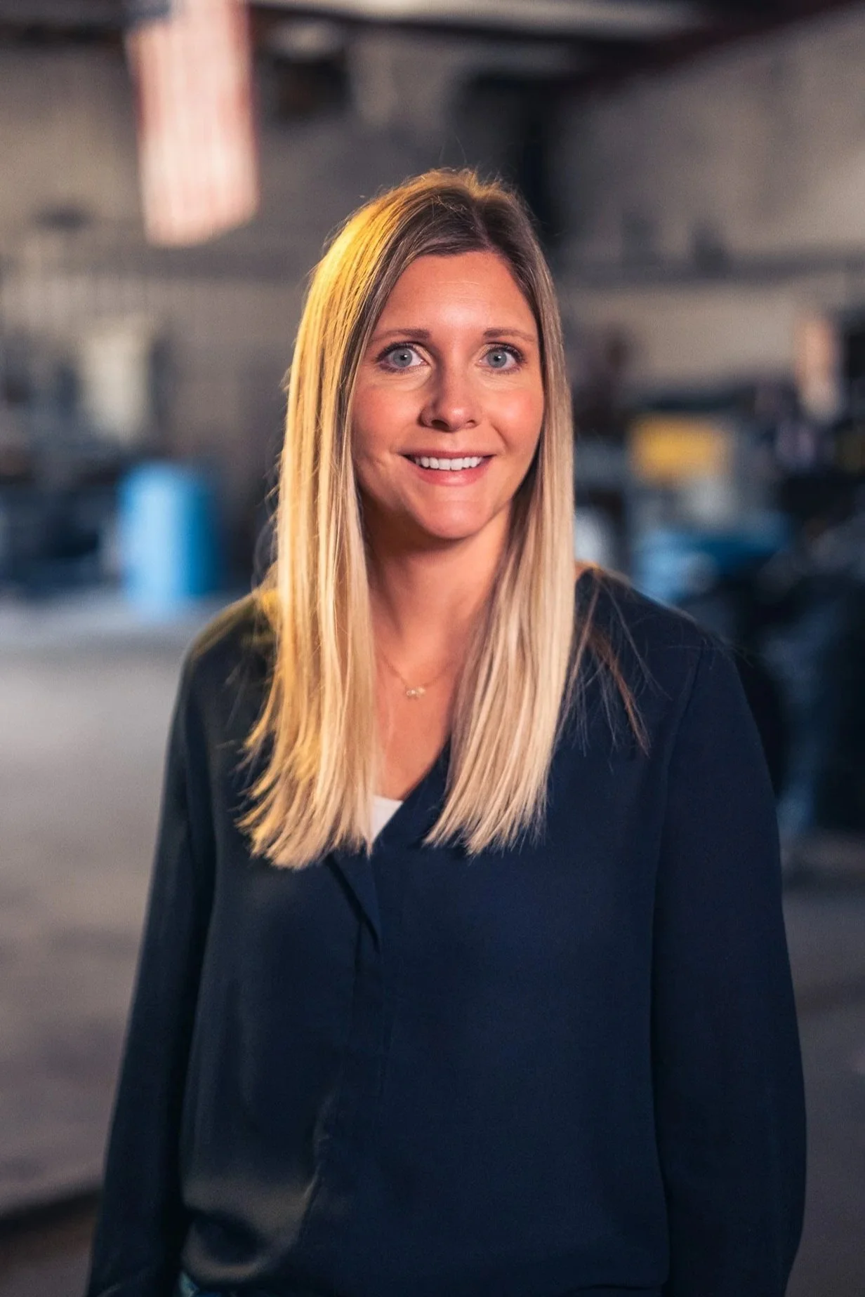 A woman with long blonde hair, blue eyes, and a friendly smile, wearing a dark blazer over a white shirt, standing in an industrial setting with blurred background.