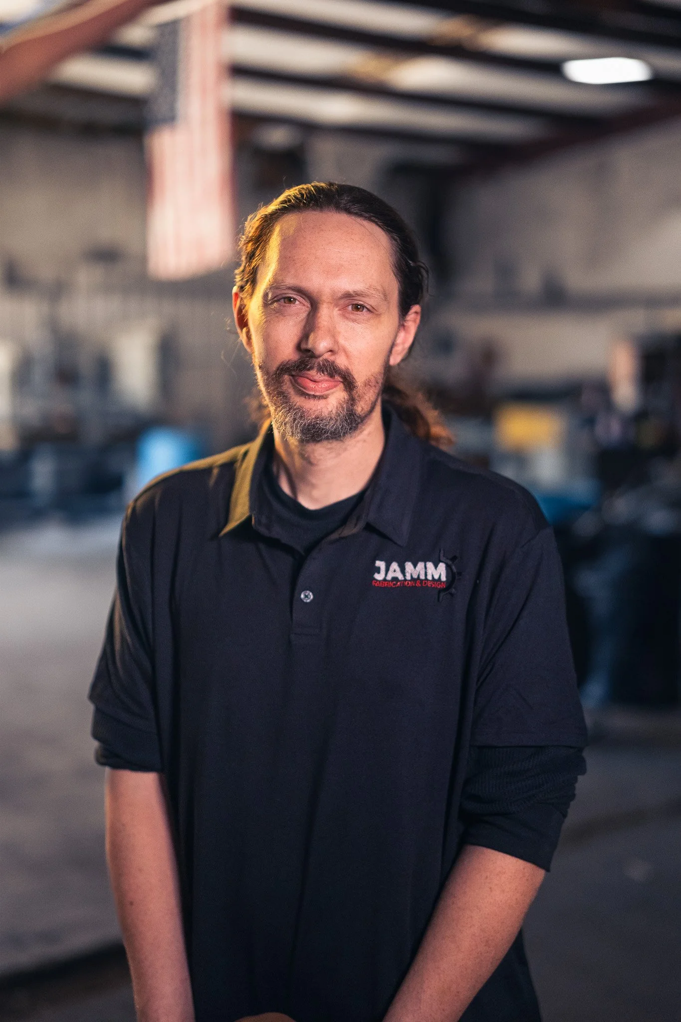 A man with long hair, a beard, and mustache, wearing a black jacket with a logo that says 'JAMM' on it, standing in a workshop or industrial space.