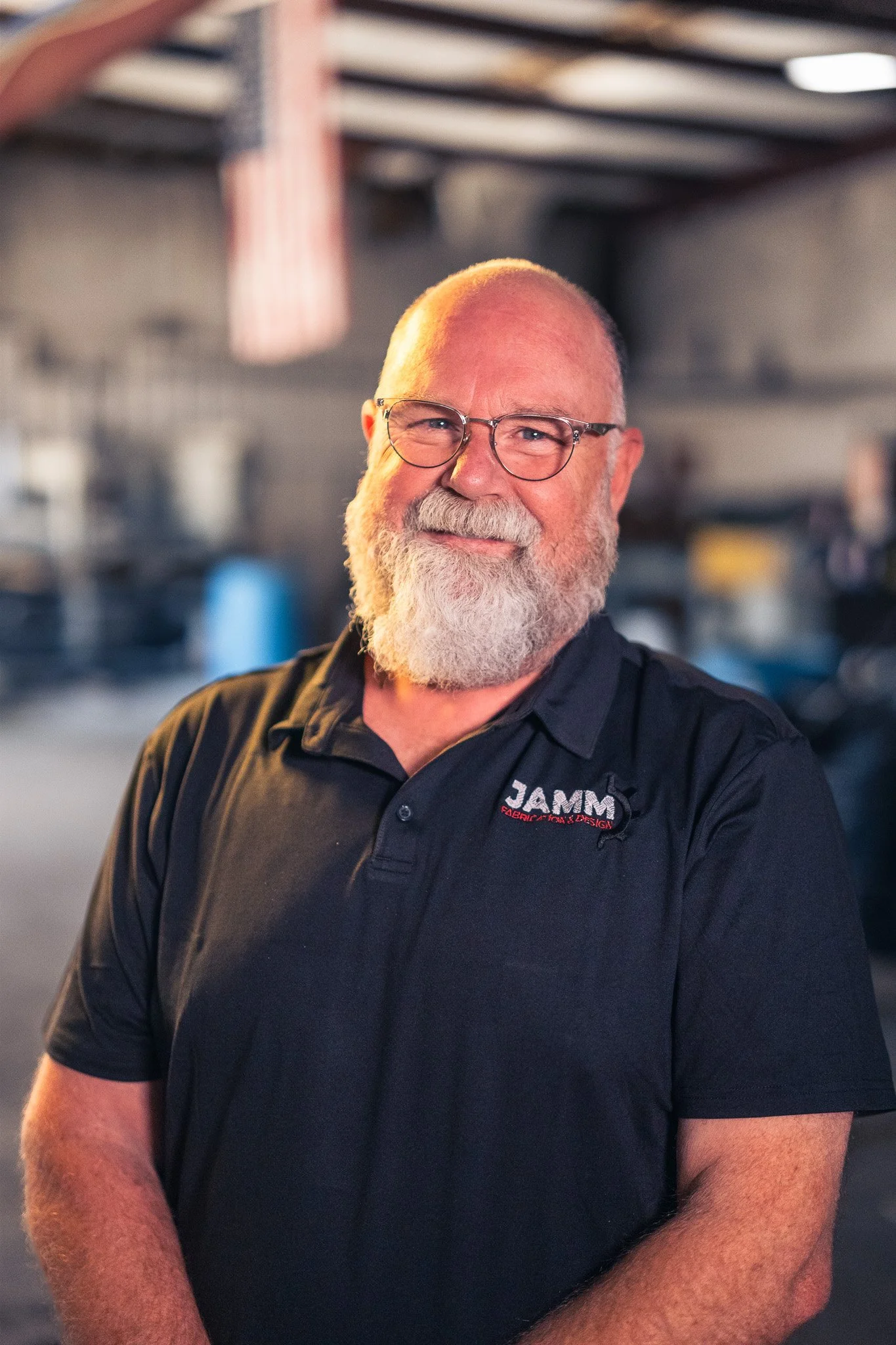 A man with glasses and a white beard, wearing a black polo shirt with a logo that reads 'JAMN Fabrication & Design,' standing inside a workshop with tools and equipment in the background.