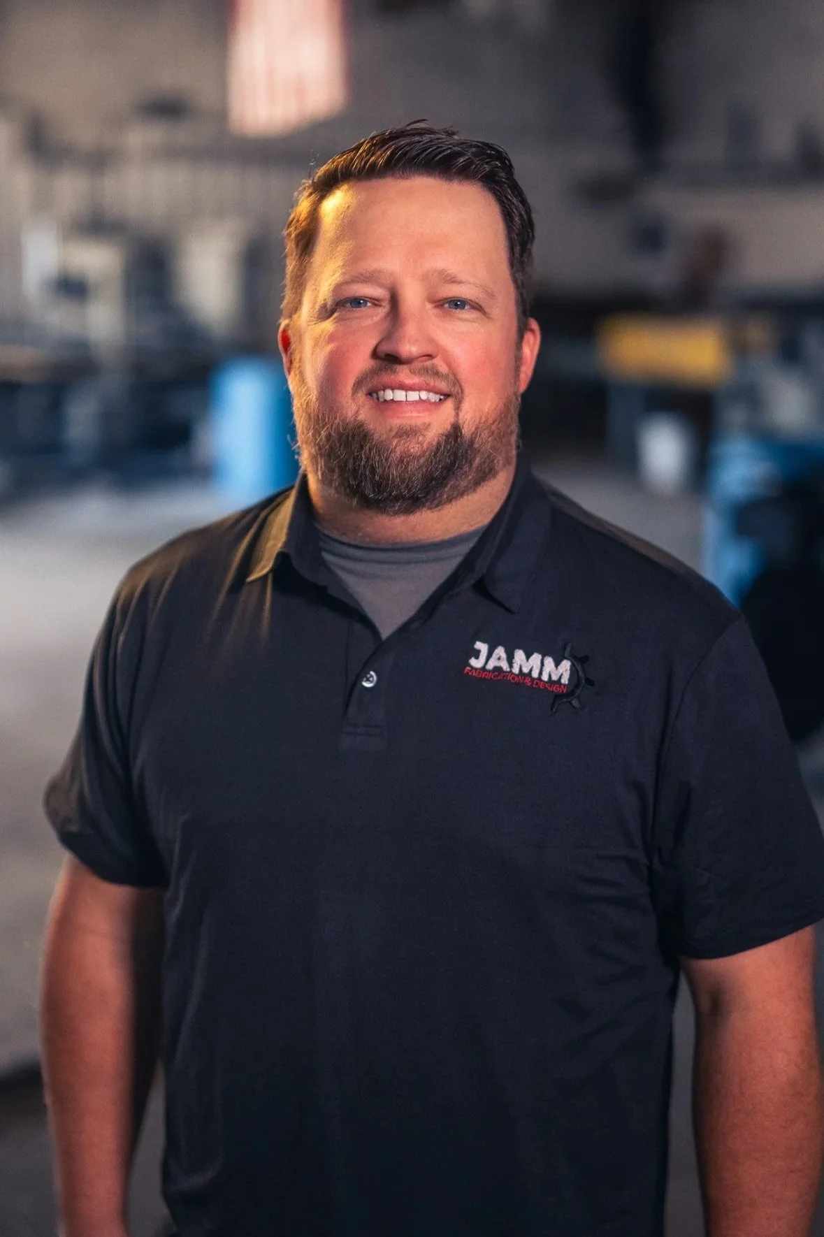 A man with a beard and short hair smiling at the camera in a workshop setting, wearing a black polo shirt with a logo that says JAMM Fabrication & Design.