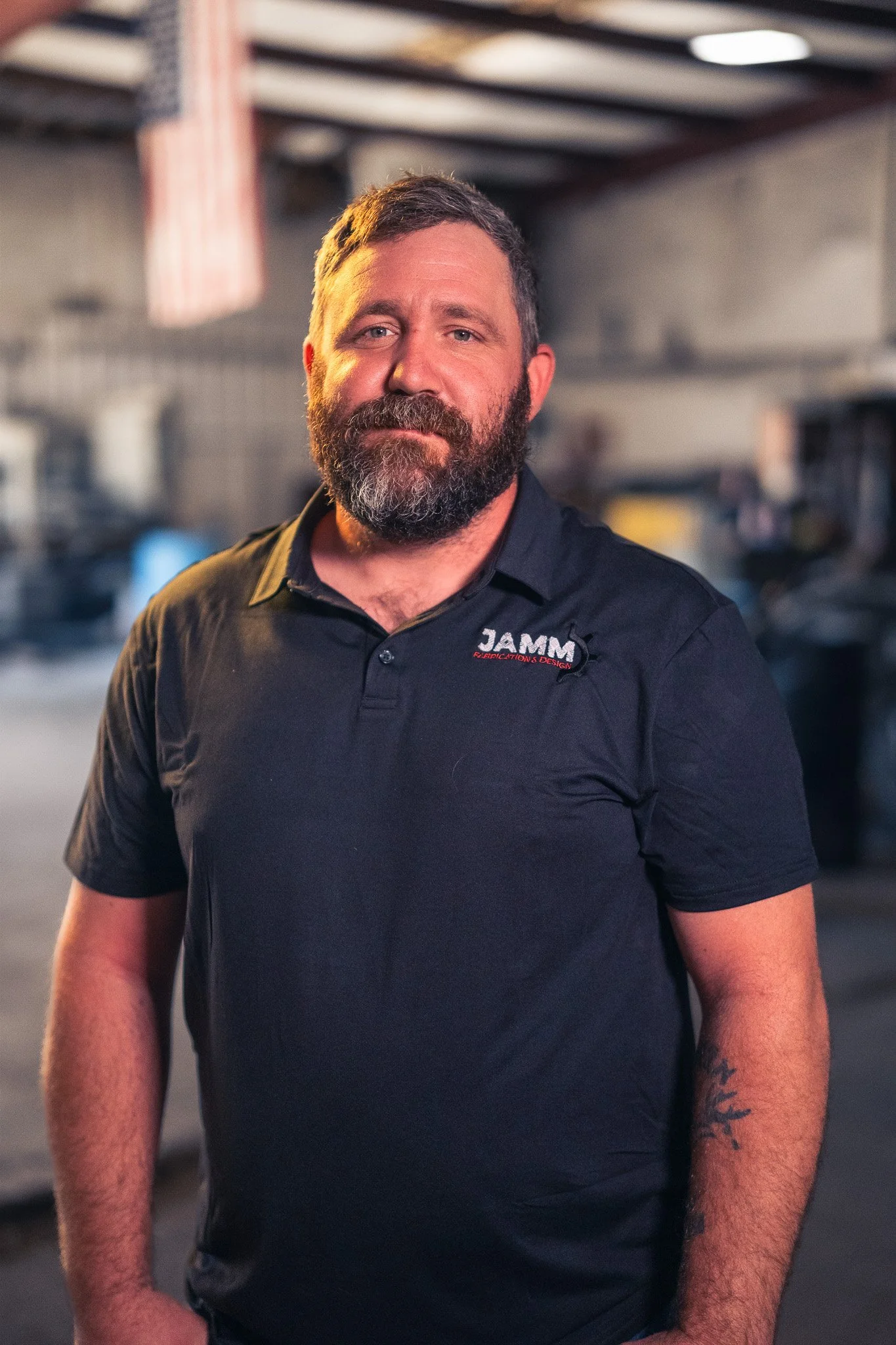 A man with a beard and short hair standing in an industrial-style setting, wearing a black polo shirt with 'JAMM' embroidered on it, smiling softly at the camera.