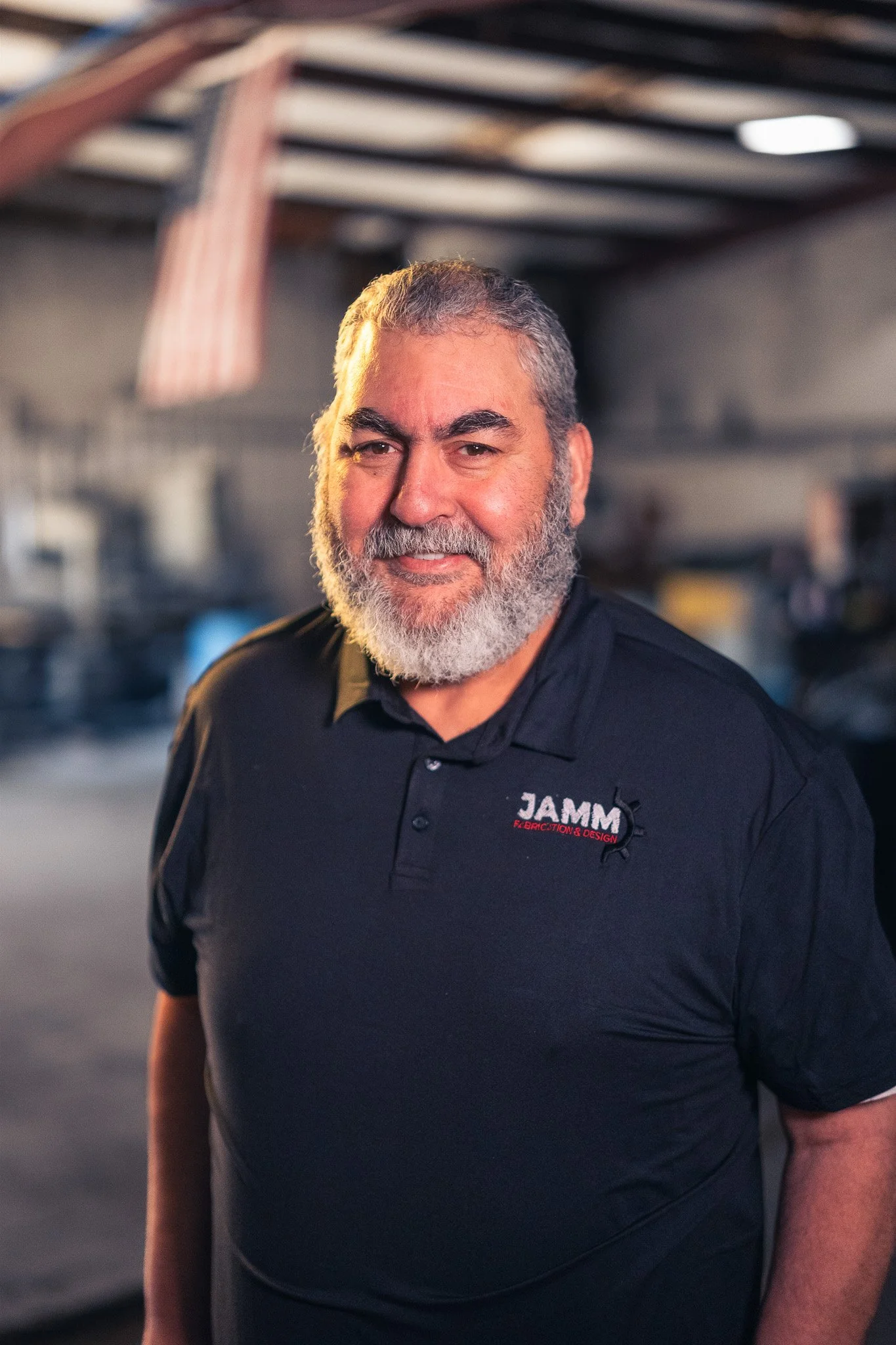 A man with a beard smiling, wearing a black polo shirt with a logo that says 'JAMM Fabrication & Design', standing inside a workshop or industrial space with blurred background.