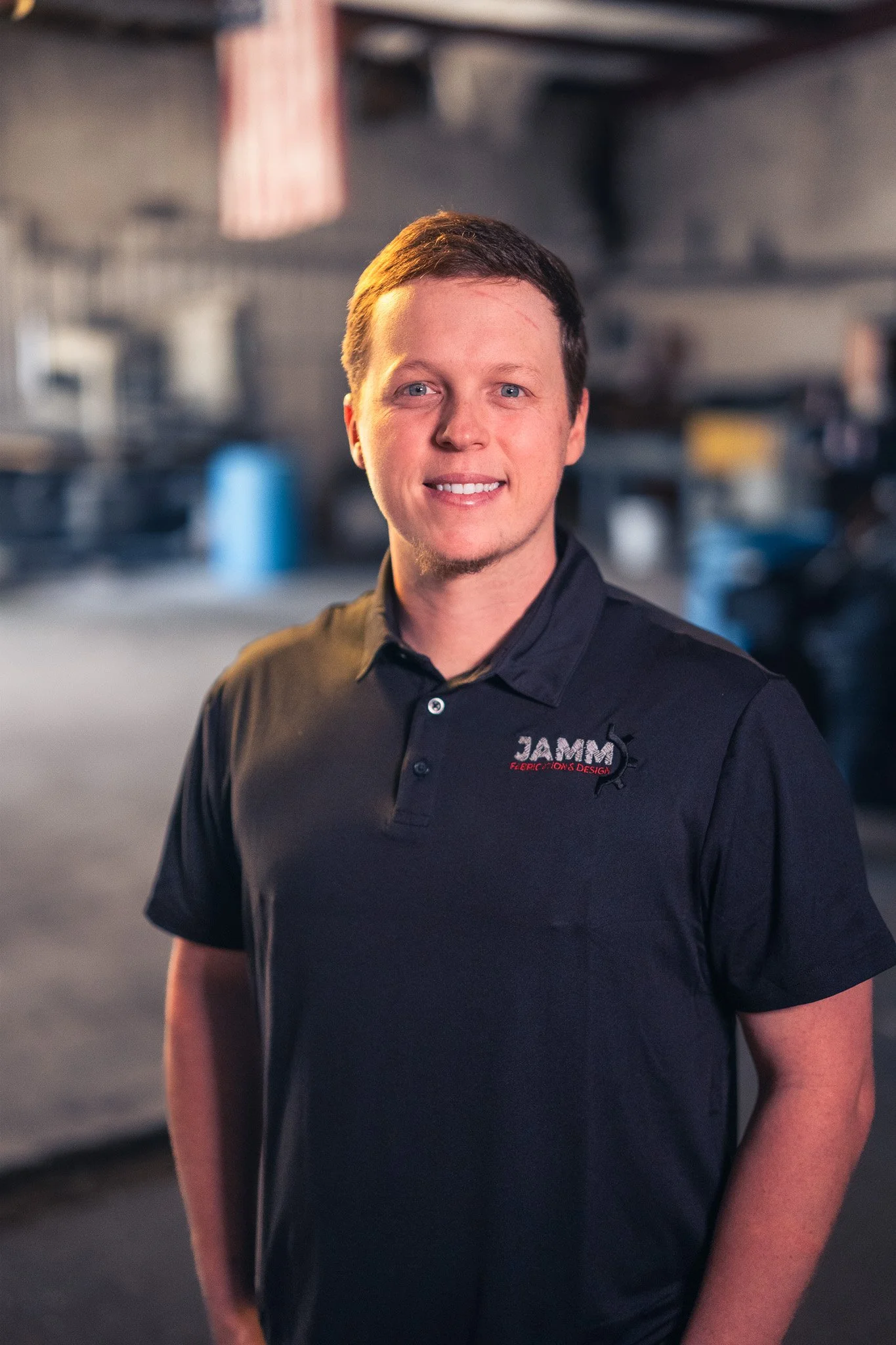 A young man with short brown hair and blue eyes, smiling, wearing a black polo shirt with a logo that says "JAMM Electric, Controls & Design" on the left chest, standing inside a workshop or industrial space with blurred tools and equipment in the background.