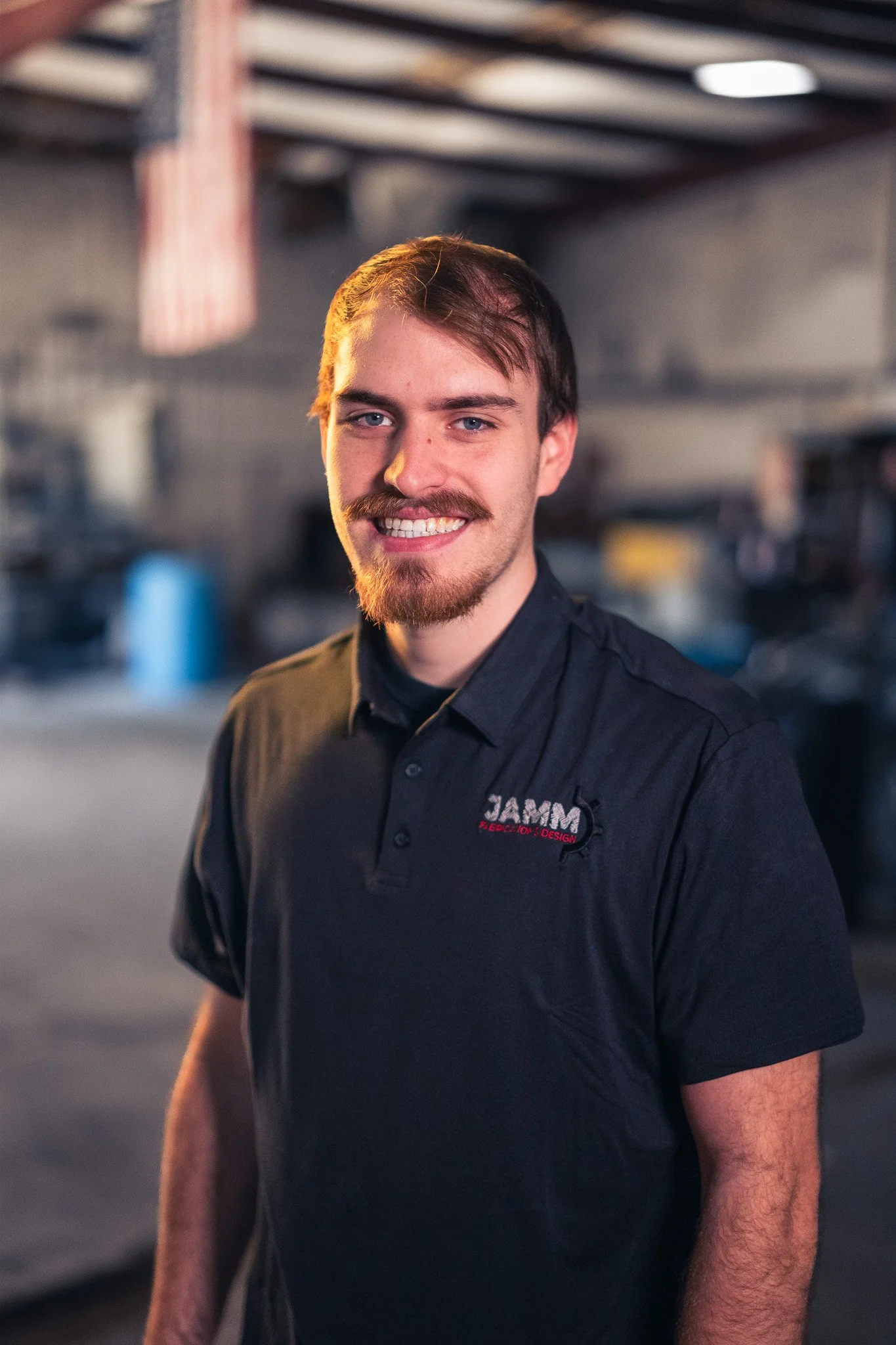 Young man with brown hair and beard wearing a black shirt standing indoors, smiling at the camera.