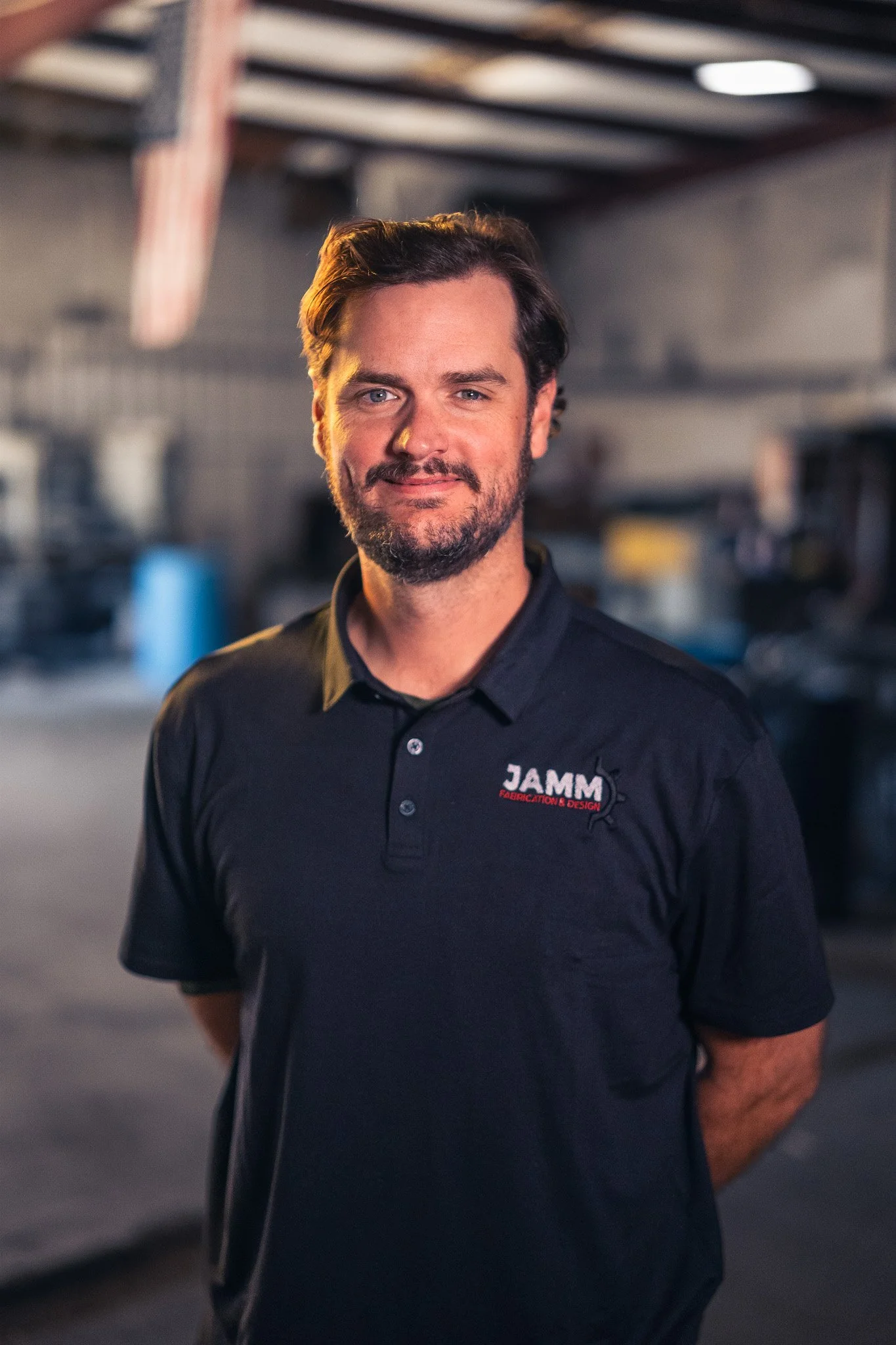A man with brown hair and a beard, wearing a black shirt with a logo that reads 'JAMM' in red and white, stands in a workshop or manufacturing setting with a slightly blurred background.