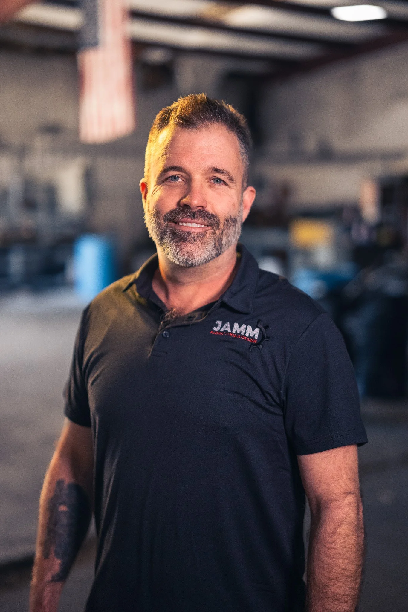 A man with gray beard and short gray hair smiling at the camera, wearing a black polo shirt with a logo. He is in a workshop or garage with tools and equipment in the background.