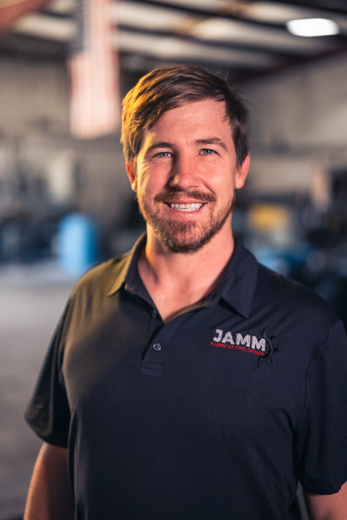 A smiling man with a beard wearing a black polo shirt with the logo 'JAMM Fabrication & Design' on it, standing in a workshop or garage setting.