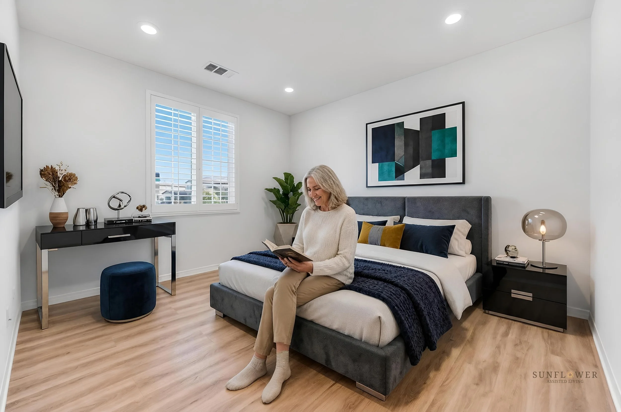 A woman sitting on a bed, smiling and reading a book in a modern bedroom with white walls, wooden floor, large window with shutters, artwork on the wall, and a bedside table with a lamp.