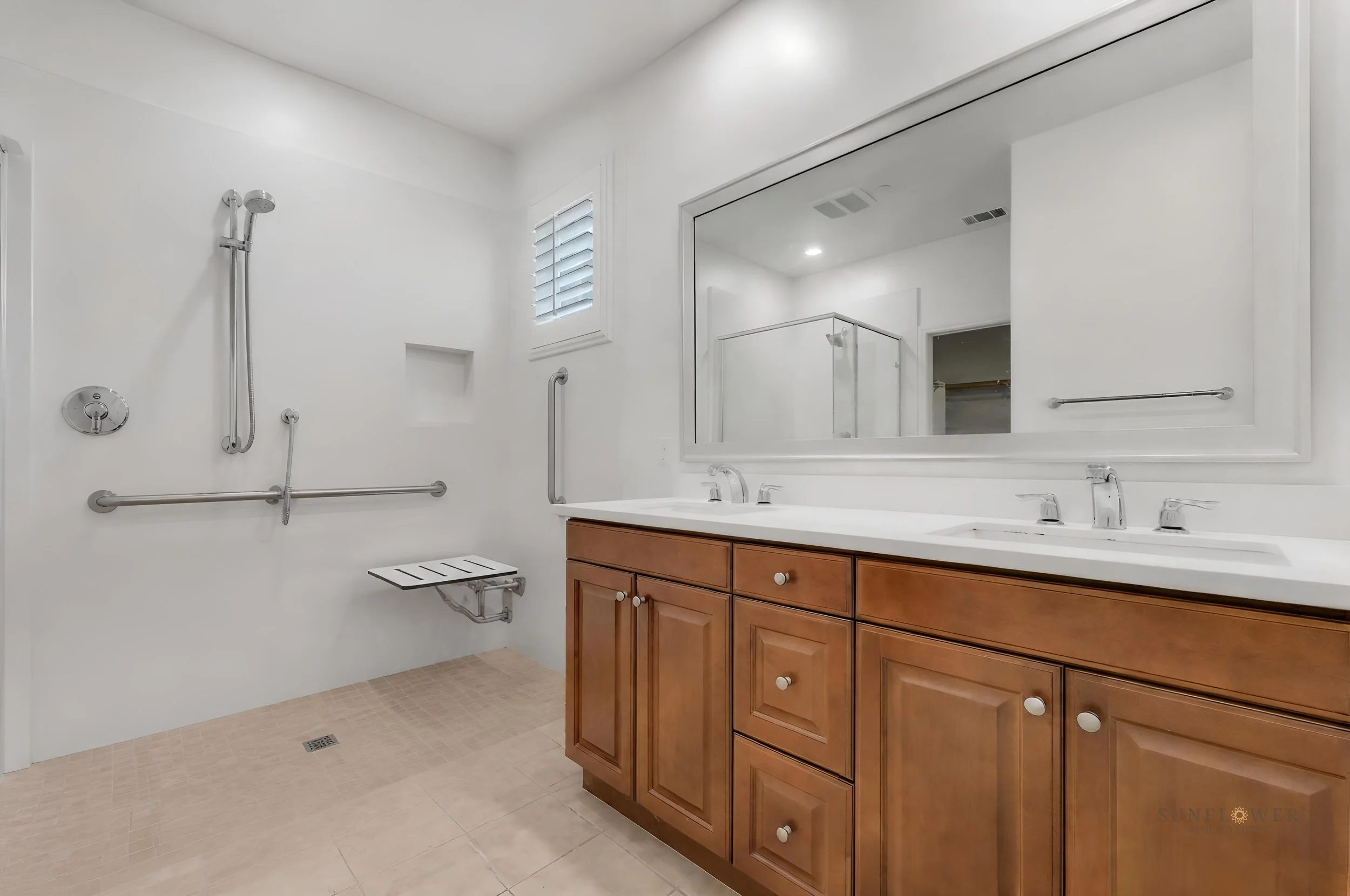 Modern bathroom with wooden vanity, double sinks, large mirror, shower area with grab bars, small window with blinds, and light-colored tiled floor and walls.