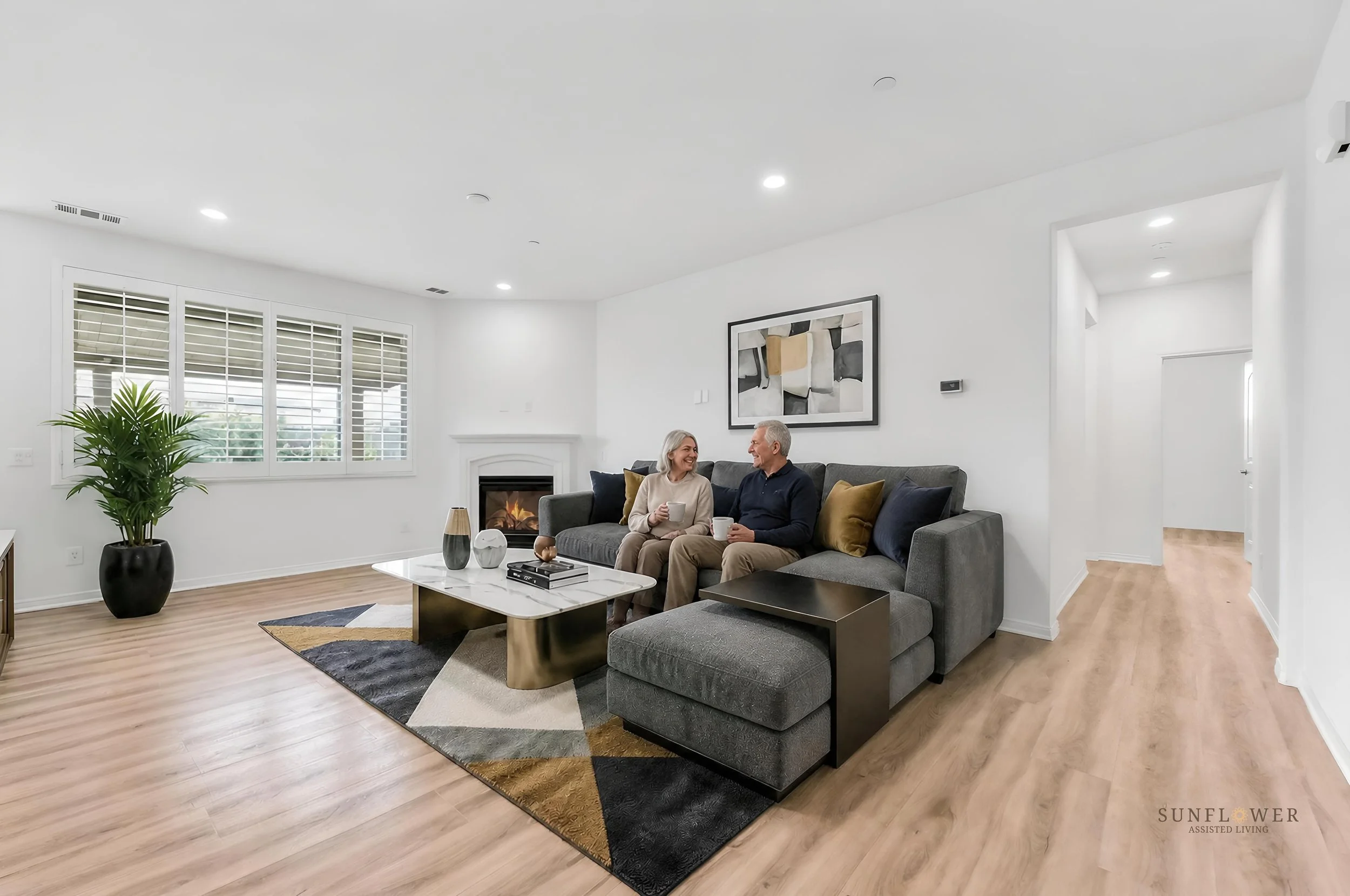 A living room with white walls, a gray couch, a coffee table, a large potted plant, abstract wall art, and two people sitting on the couch holding coffee mugs, smiling at each other.