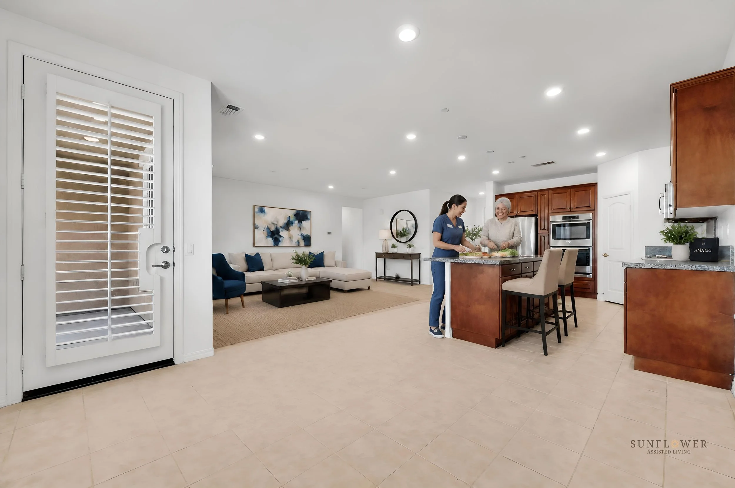 Interior of a living room and kitchen with two women preparing food at a kitchen island. The living room has a beige sofa with blue pillows, a coffee table, and a painting on the wall. The kitchen features wooden cabinets and stainless steel applianc