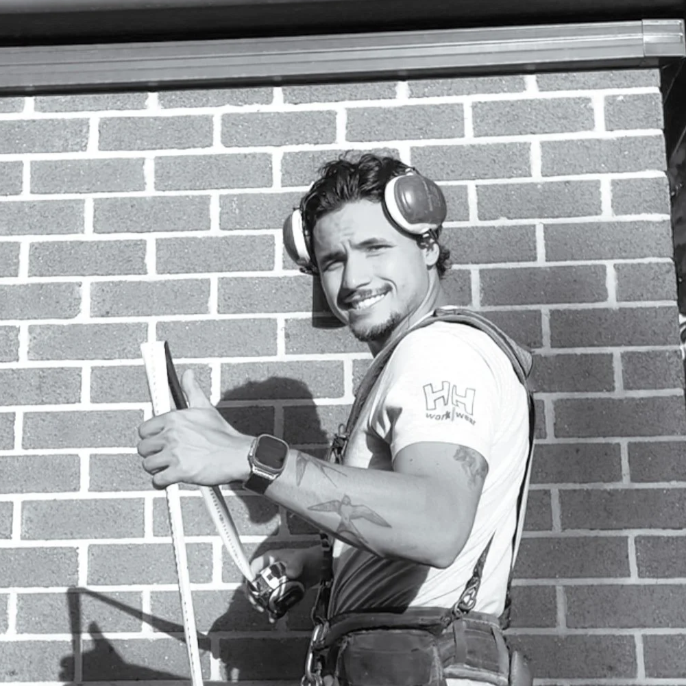 A man with headphones, a wristwatch, and a tattoo on his arm, smiling and giving a thumbs-up while holding a rope, standing against a brick wall.