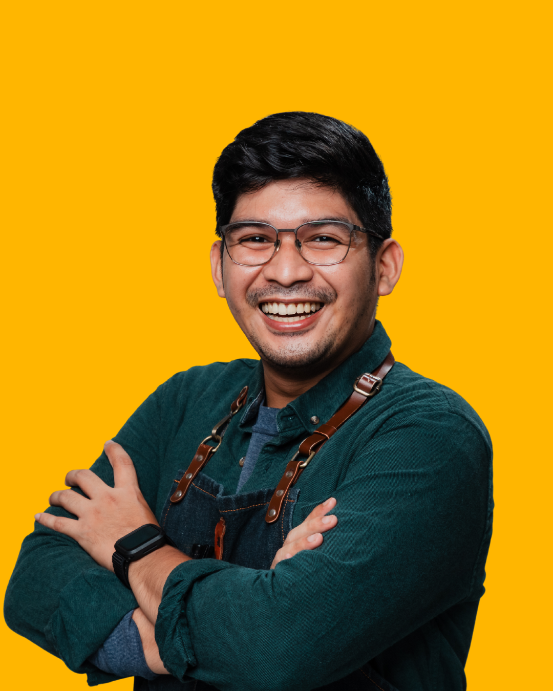 A smiling man with glasses, wearing a dark green shirt and apron, standing with arms crossed against a yellow background.