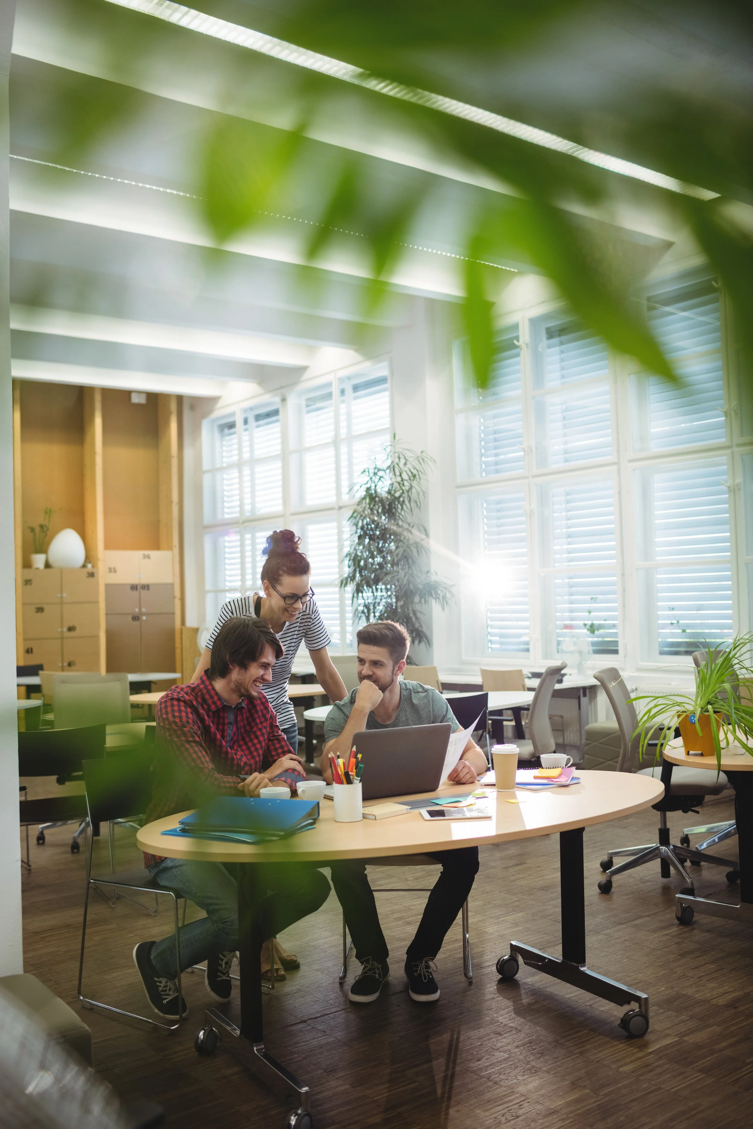 Three coworkers gathered around a desk looking at a laptop and papers, smiling and sharing a laugh in a bright, modern office with large windows and indoor plants.