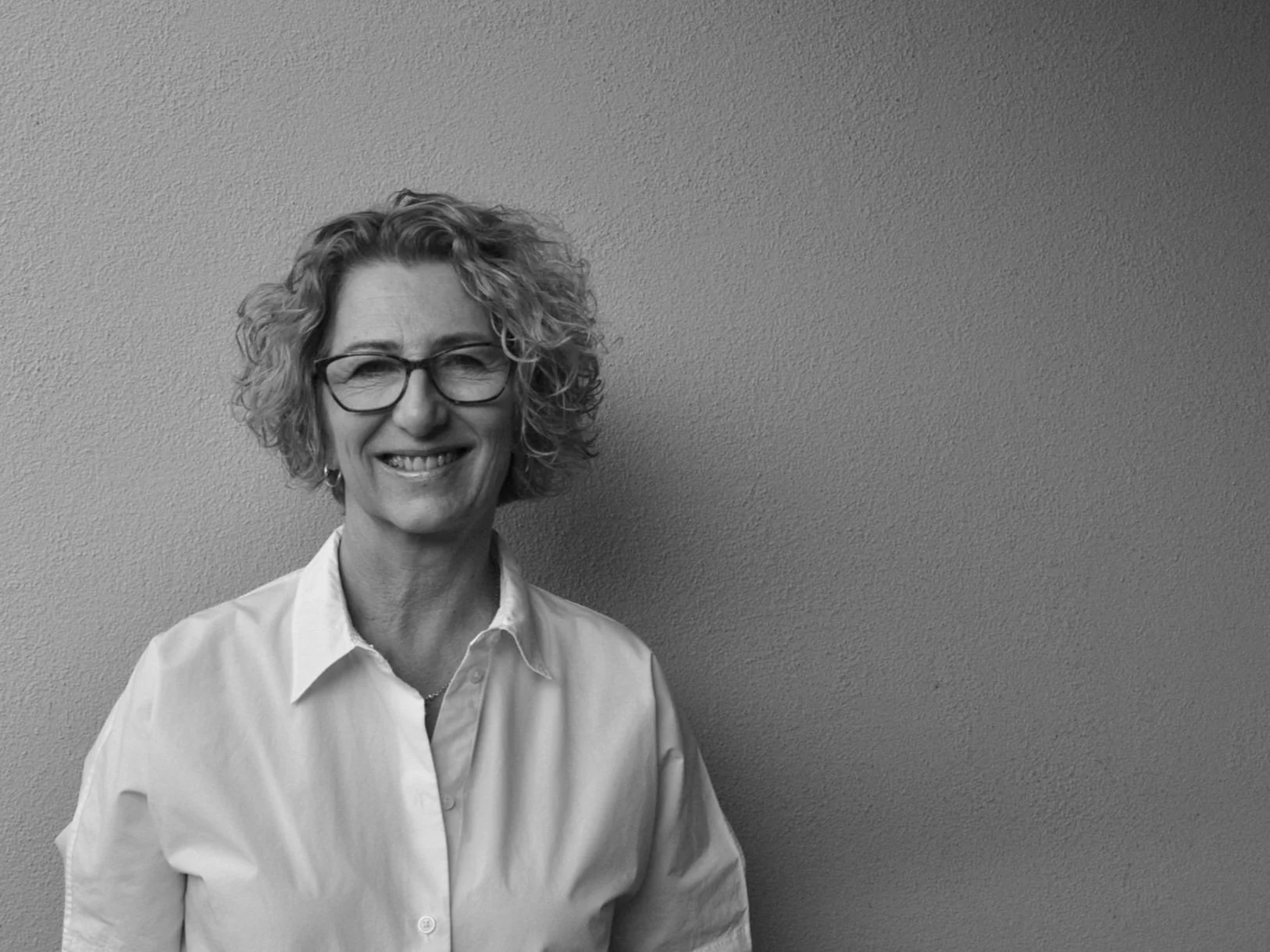 Black and white portrait of a woman with curly hair, wearing glasses and a white shirt, smiling against a plain wall.