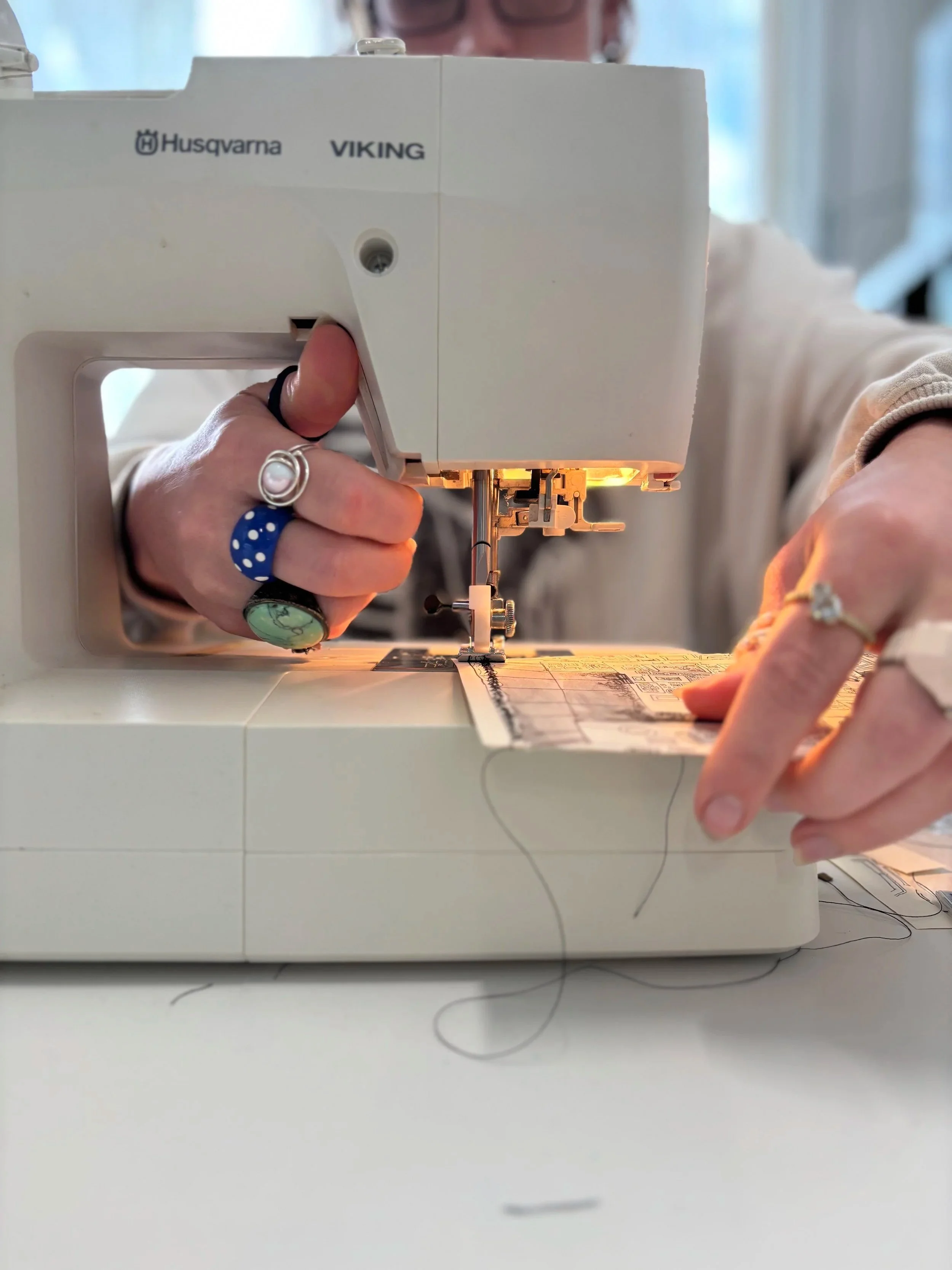Close up of the foot of a sewing machine sewing paper, with two hands wearing colourful and patterned rings
