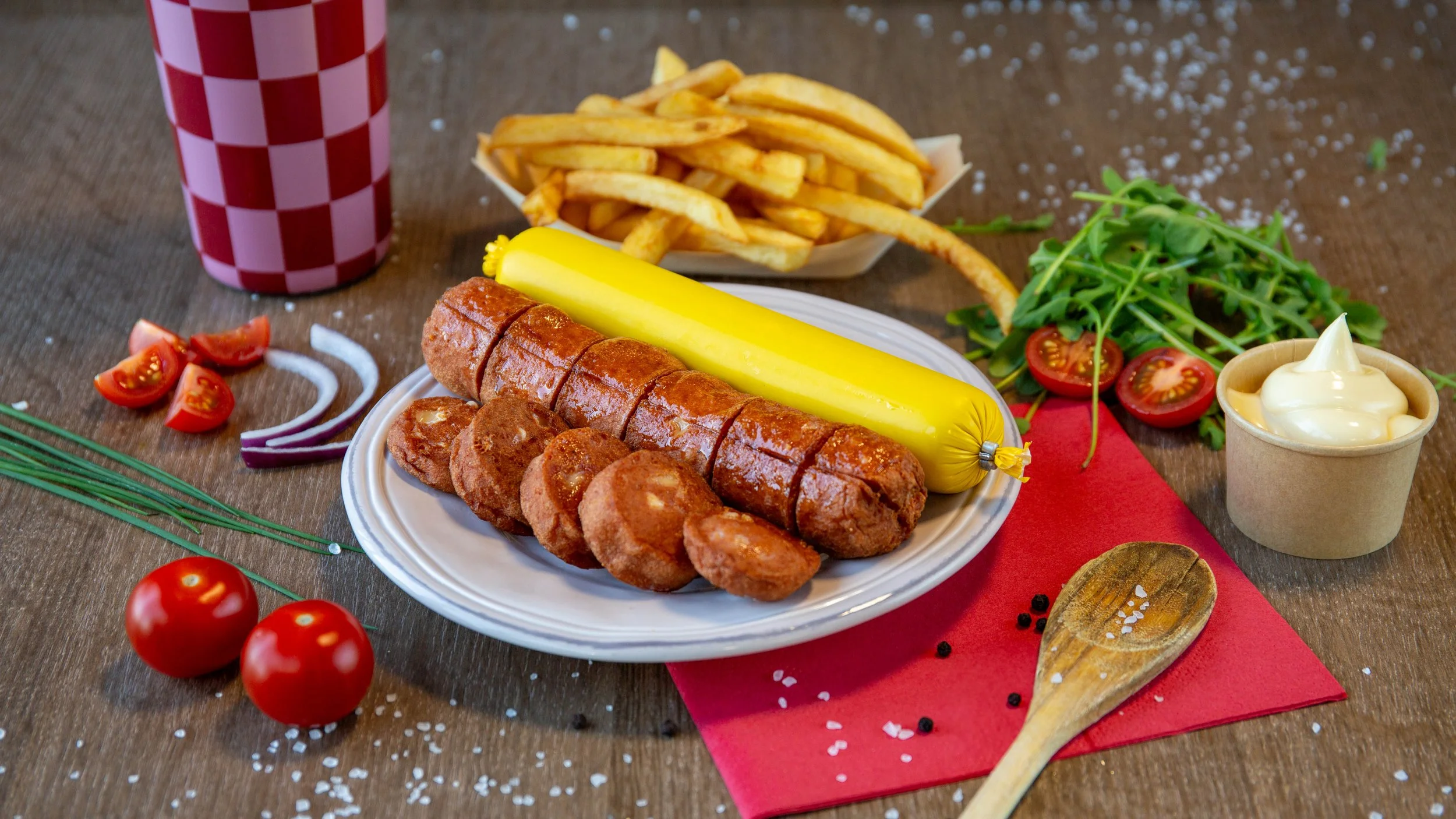 Foto van een bord met gegrild vlees, een worst, een kom frietjes, tomaten, kaas, salade en een glas met een rood-wit geruit patroon.