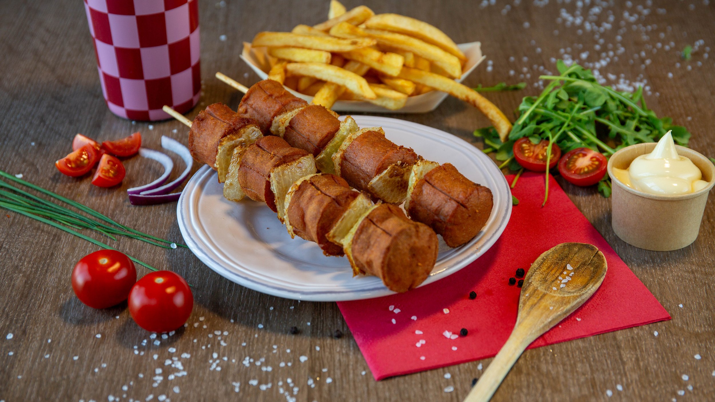 Gefrituurde worstjes op een wit bord, met gefrituurde patat en een glas frisdrank met rood-wit geruit patroon, omringd door tomaatjes, sla, uien, en container met mayo op houten tafel.