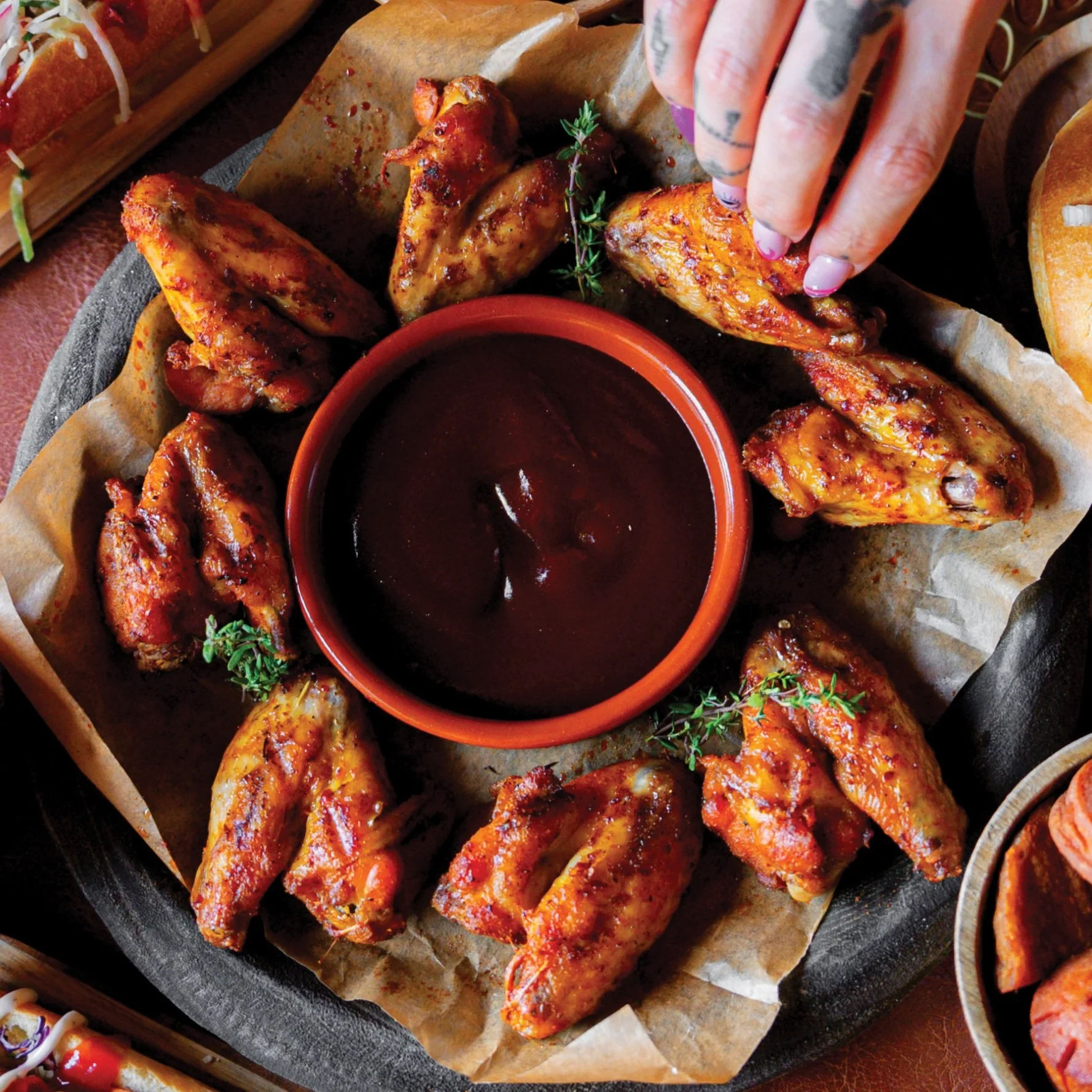 Gouden kippenvleugels met een zwarte barbecuesaus in een oranje kom, versierd met tijmblaadjes, geserveerd op een papieren ondergrond.