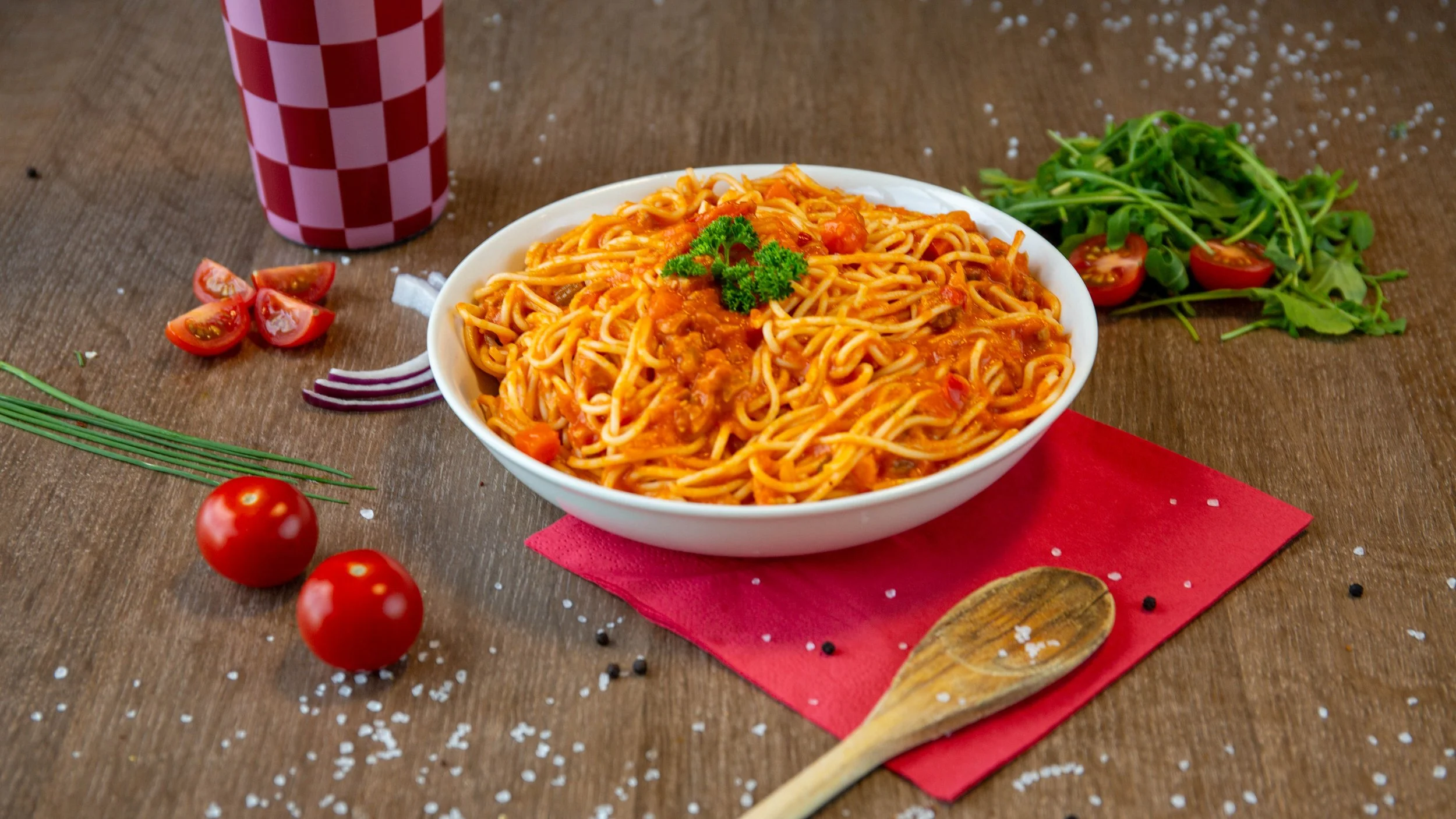 Een kom met spaghetti bastaard met tomatensaus, omringd door cherry tomaten, rode ui, basilicum, rucola, en een drankje met een rood-wit geruit patroon.