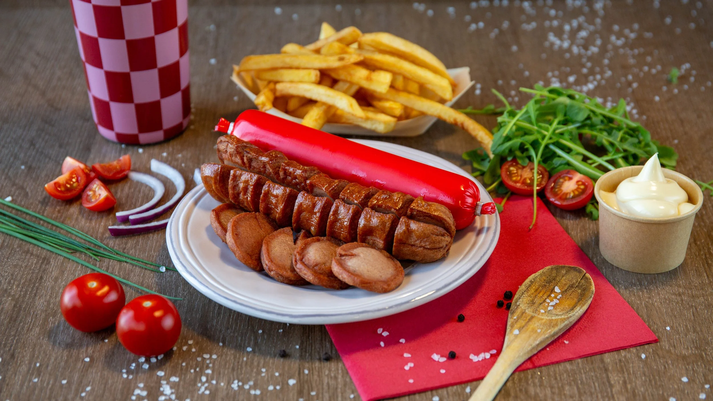 Een bord met worst, een hotdog, friet, cherrytomaten, sla, een beetje ketchup en mayonnaise op een houten tafel.