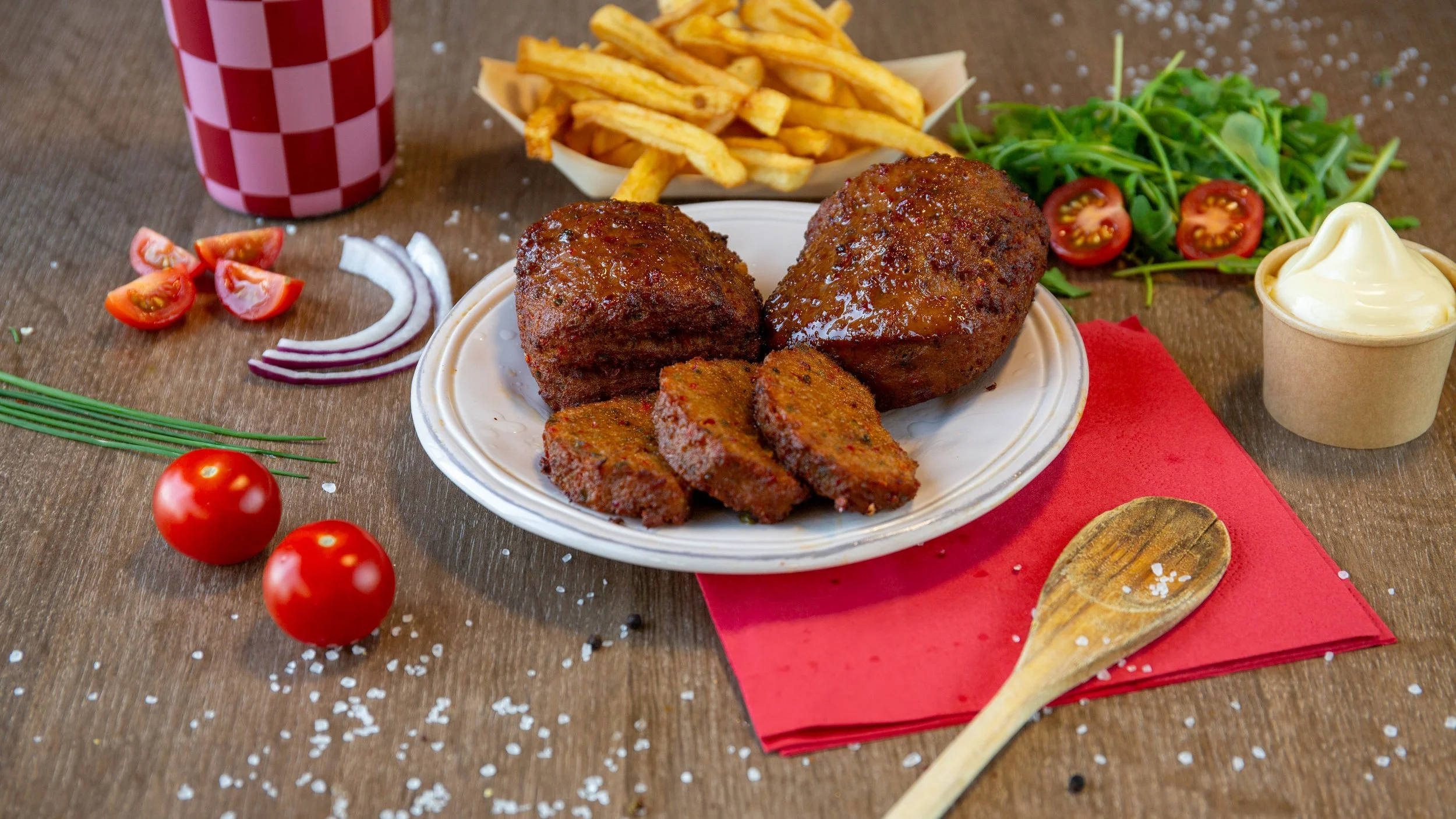 Gerecht met twee grote gehaktballen en plakjes vlees, geserveerd met frietjes, salade met tomaat en radijs, en mayosoep, op een houten tafel.