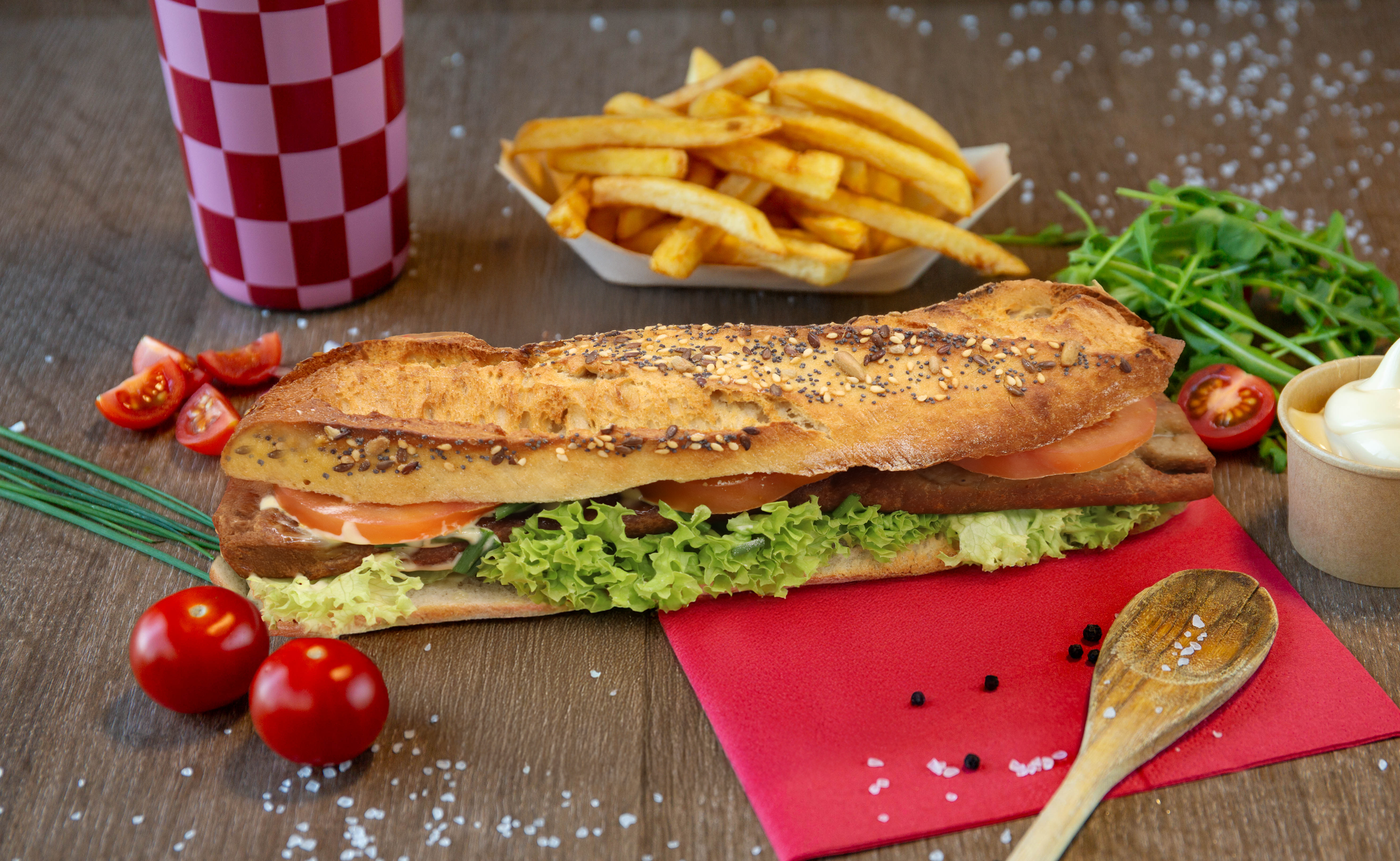 Een finetegere baguette met sla, tomaat, ham en smeerkaas, omringd door cherrytomaten, frieten, een frisdrank, salade met waterkers, en een mengsel van peper en zout, op een houten tafel.