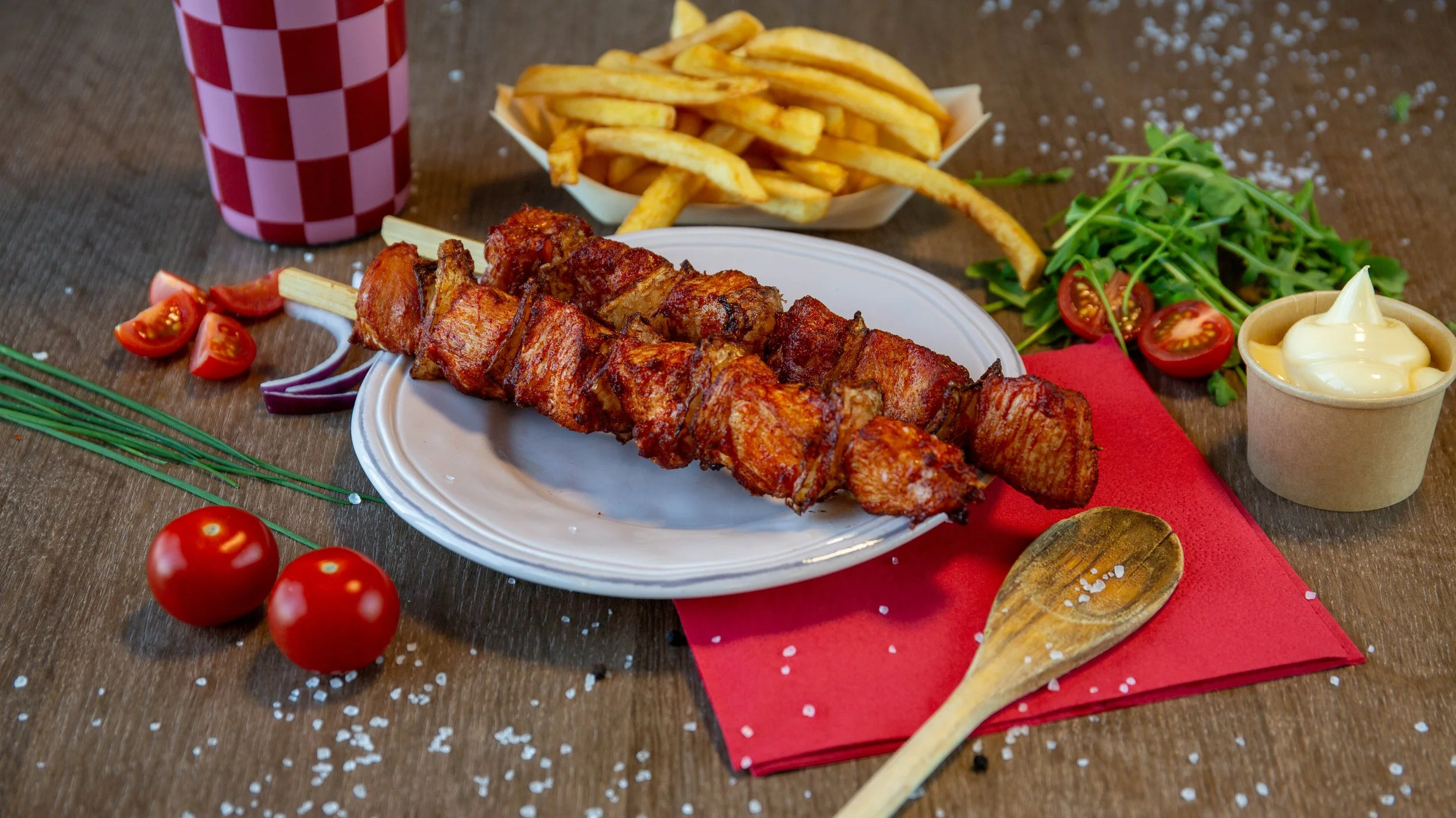 Gemarineerde kipspies op een wit bord met frietjes, cherrytomaten, komkommer, ringen rode ui, een klein bakje mayonaise, en een glaasje met een rode en witte ruitjespatroon op de achtergrond.