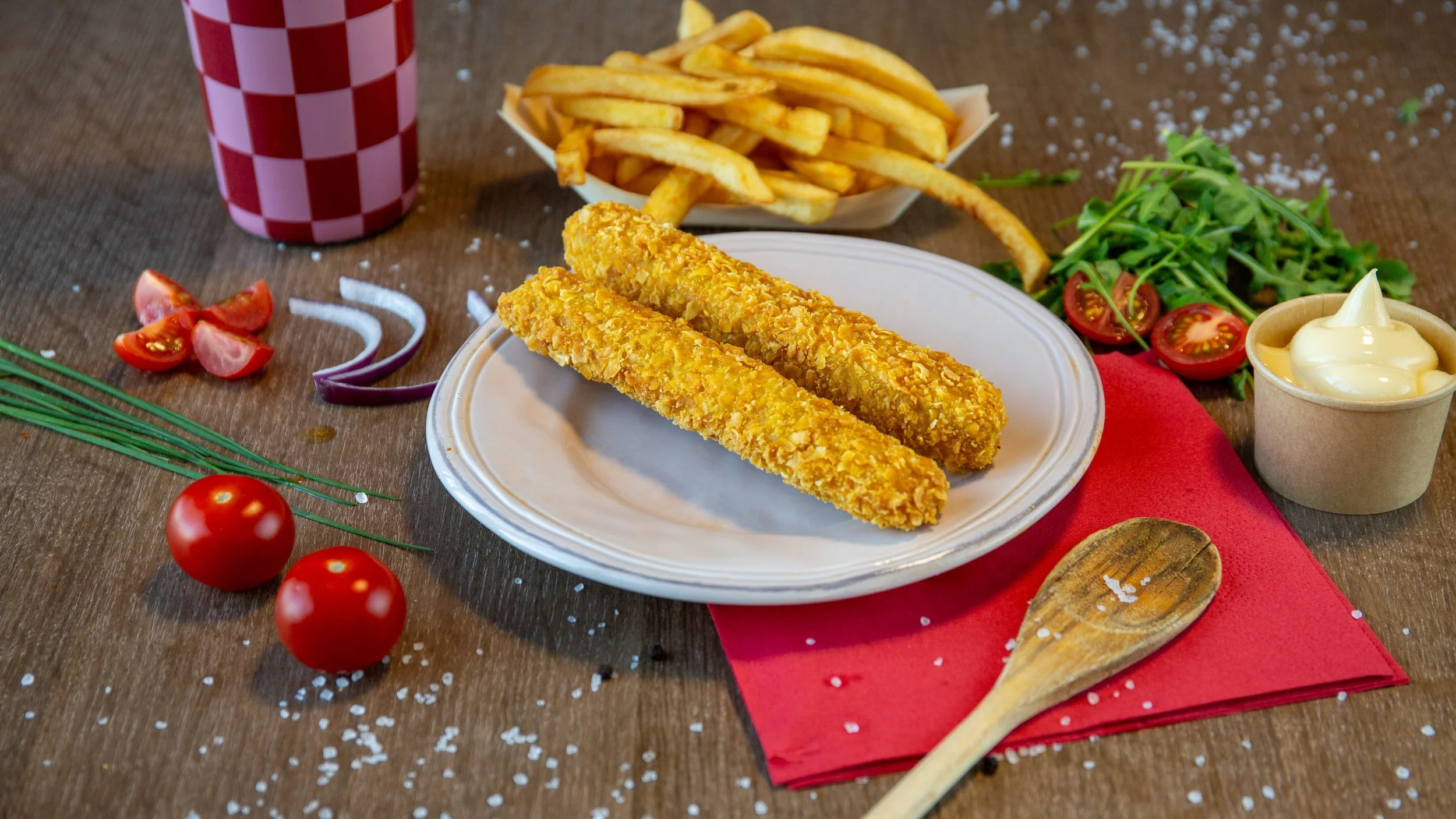 Gefrituurde vissticks met frietjes, tomaten, cherrytomaten, salade, mayonaise, en frisdrank op tafel.