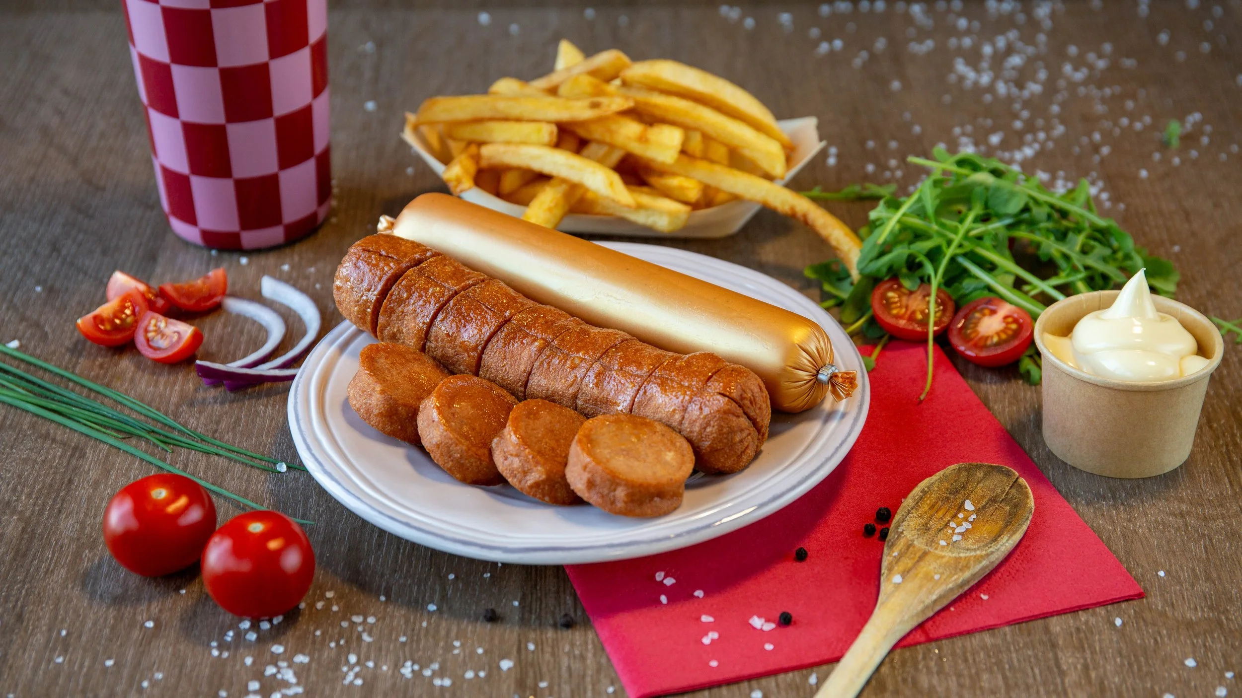 Een bord met frieten, een hotdog, kroketten, en een kaasworst, naast een glas frisdrank, tomaten, sla, uien, en mayonaise op een tafel.