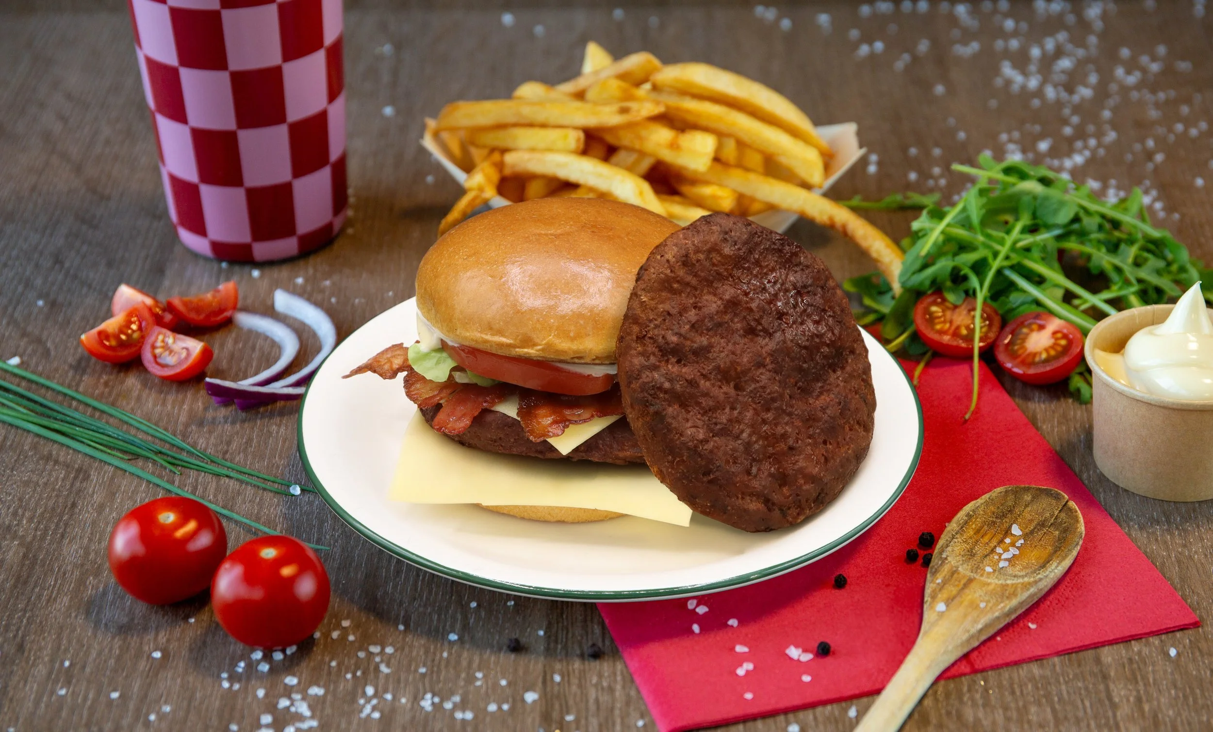 Een bord met een burger met bacon, kaas, tomaat en een vegaburger, naast Frans friet, een glas met een rood-wit geruit patroon, cherrytomaten, een salade met tomaat en rucola, en een schaaltje met mayonaise op een houten tafel.