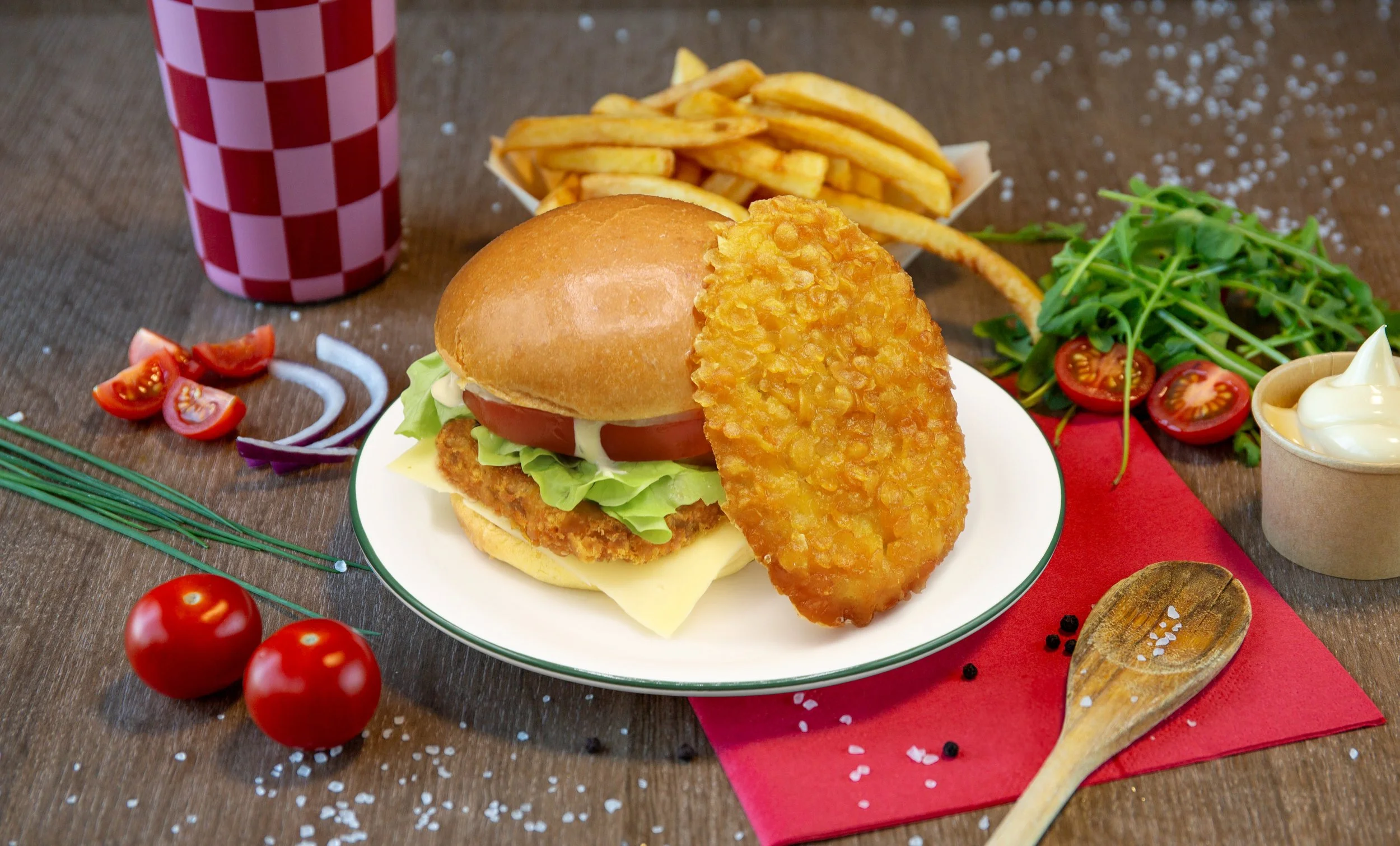 Hamburger met kaas, tomaat, sla en tartaar, friet, frisdrank, salade, cherrytomaten, rode ui, zout, peper, en mayonaise op tafel.