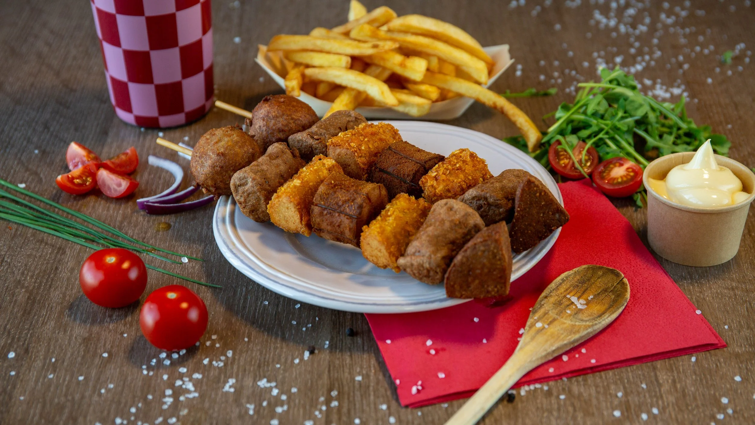 Een bord met gefrituurde bitterballen en kroketten, omringd door tomaten, sla, ui, frietjes, een beker frisdrank en een bakje mayonaise. De tafel is versierd met een rode servet en er liggen cherrytomaten en zoutkristallen.