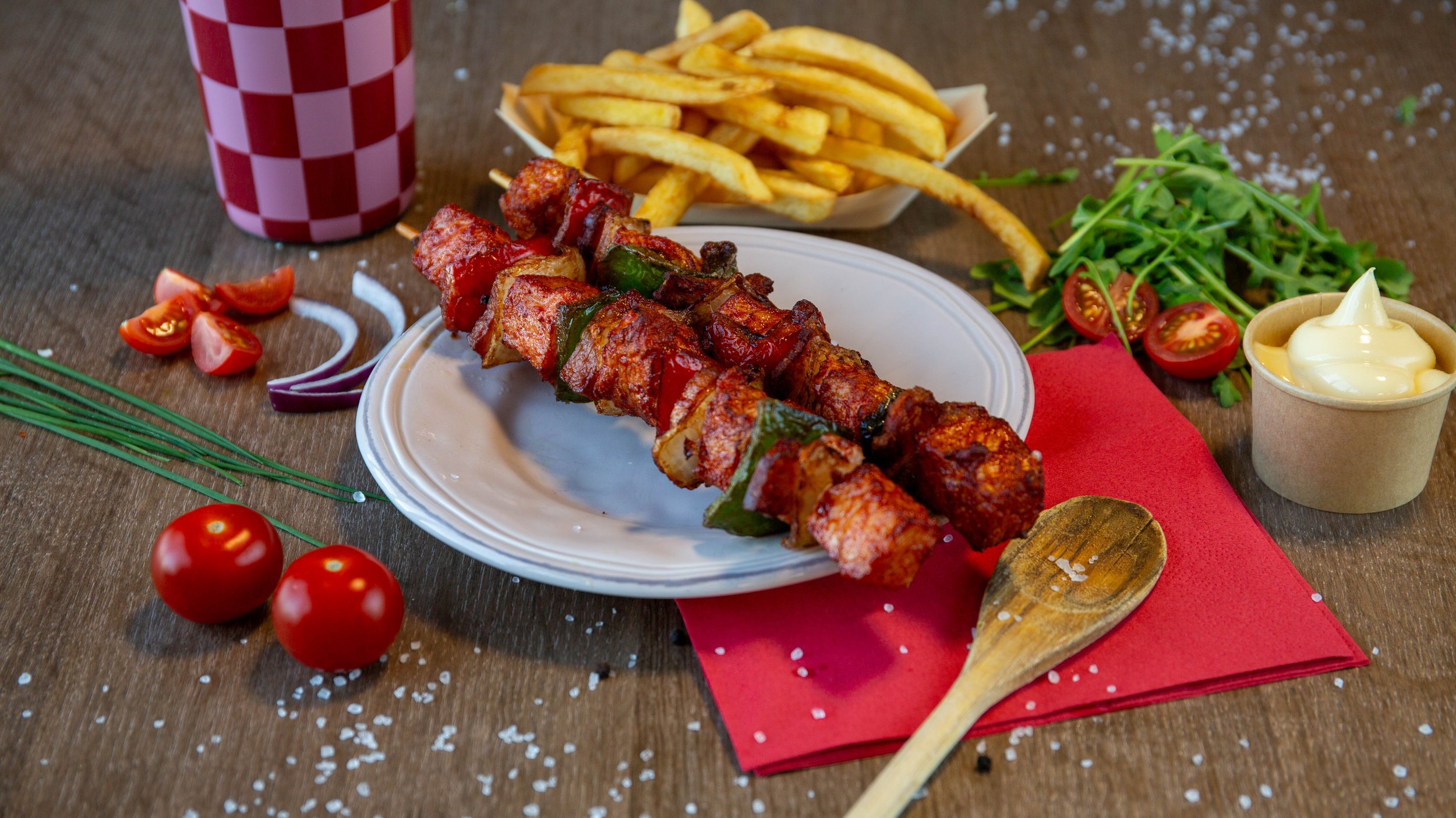 Satéspies met groenten op een wit bord, met frietjes en salade op de achtergrond, naast een beker cola en een knoflooksausje, op een houten tafel met een rode servet en gemorste zoutkristallen.