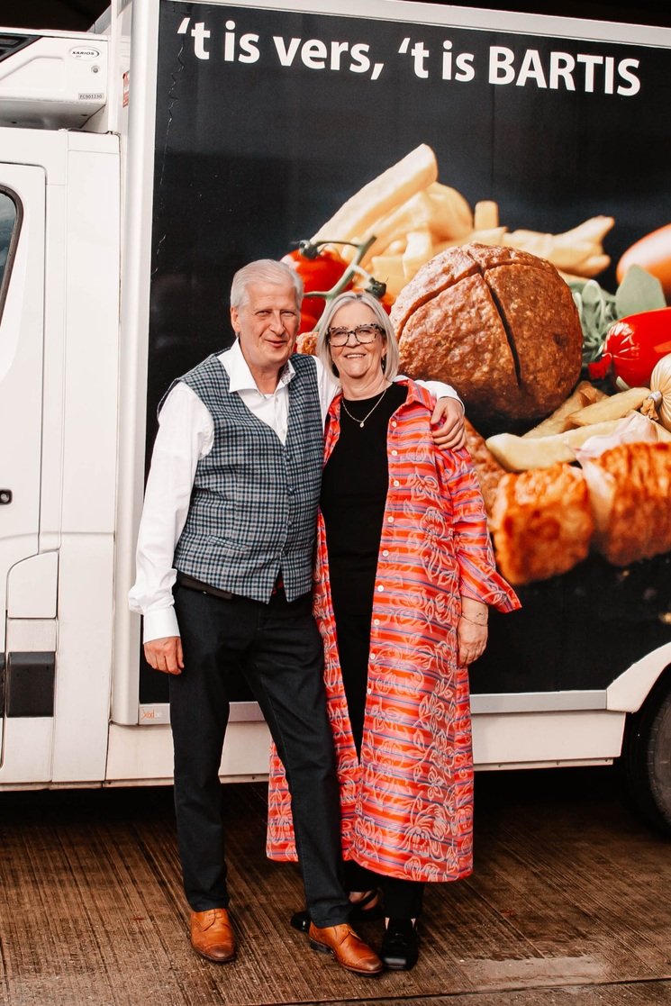 Twee mensen staan naast elkaar voor een grote vrachtwagen met een advertentie voor 't is vers, 't is BARTIS, en afbeeldingen van food, met een man in een wit shirt en een grijze vest en een vrouw in een zwart shirt met een lange, rode en oranje kimono-achtig jas en een bril.