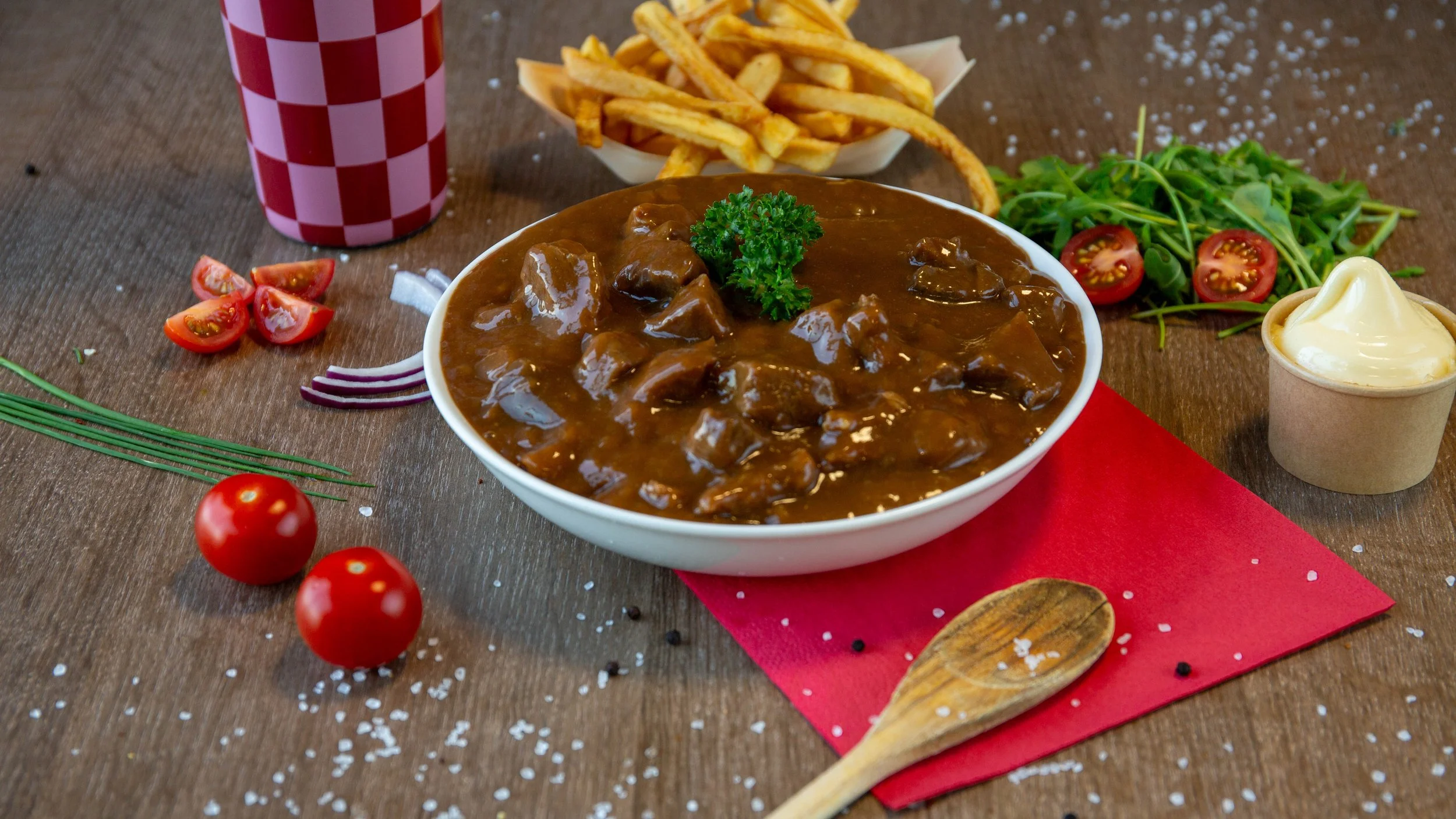 Een kom met rundvleessap met rundvlees, geserveerd met een sprietje peterselie. Omringd door cherrytomaten, gesneden rode ui, een bakje frietjes, groene salade met cherrytomaten, een beker frisdrank, en een bakje met mayonaise. De tafel is van hout en er ligt een rode placemat onder de kom.