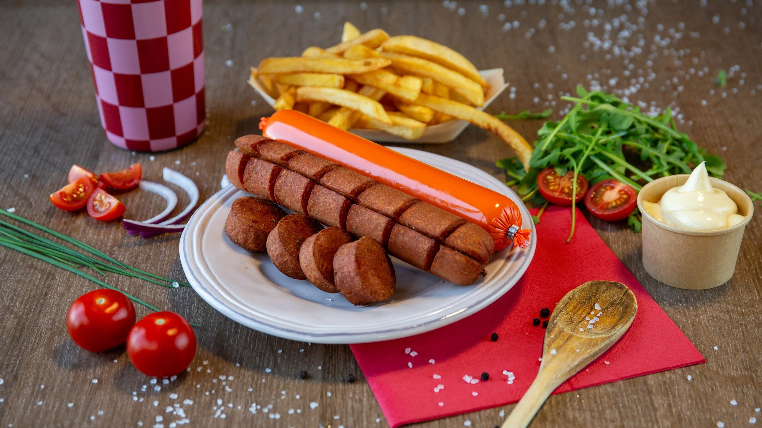 Een bord met verschillende soorten vlees, waaronder een grote hotdog en gehaktballen, met een glas soda, frietjes, tomaten, sla, uien, en een bakje mayonaise op een houten tafel.
