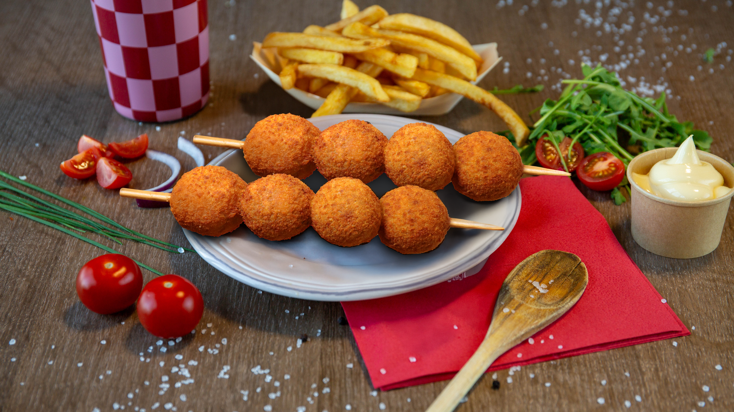 bord met frietjes, gehaktballetjes op een stokje, salades, cherry tomaten, mayonaise, een knoflooksaus in een bekertje, een glas met een rood-wit geruit patroon, een houten lepel, een rode placemat, en wat zoutkristallen op een houten tafel.
