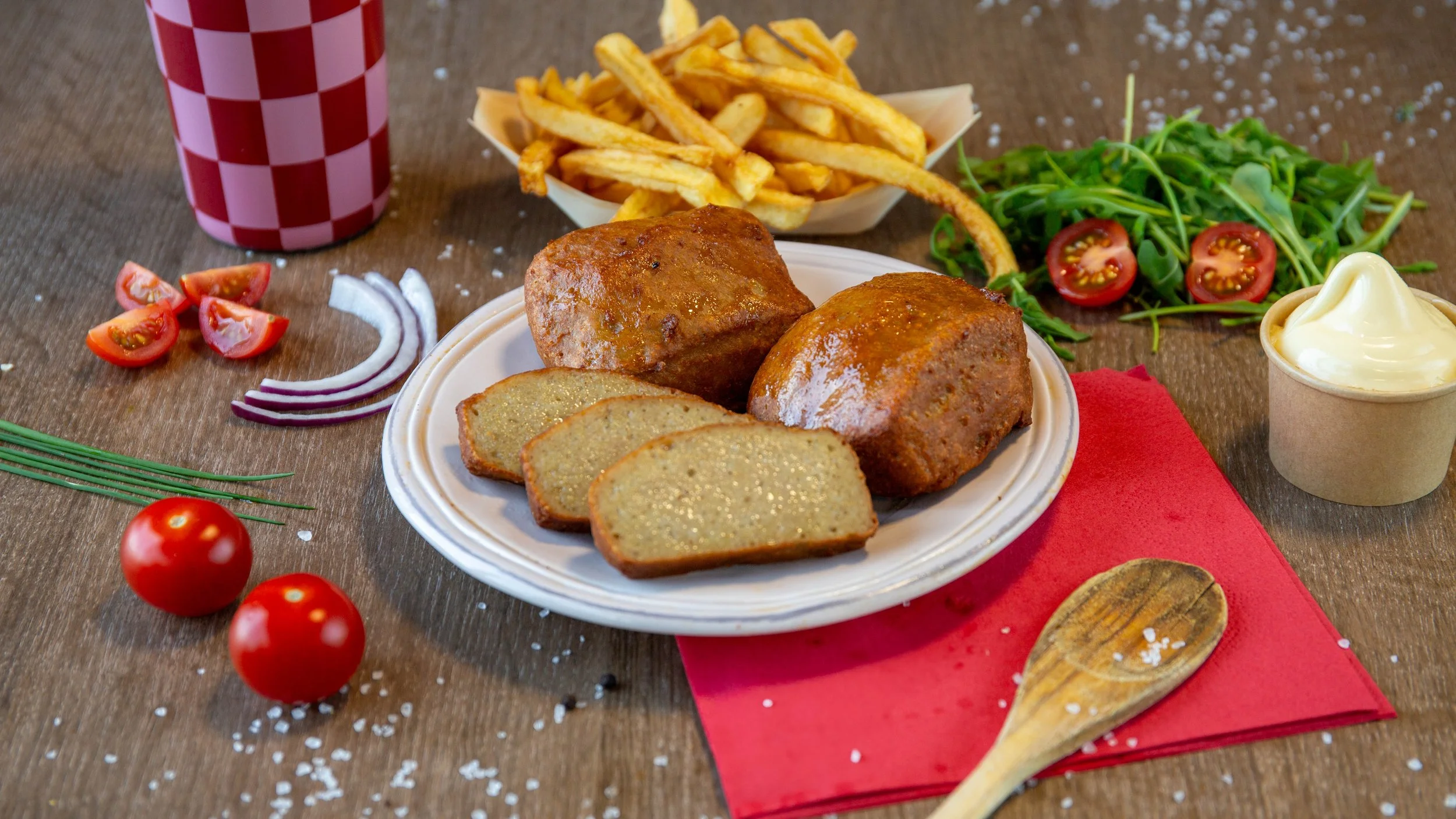 Een bord met twee gerechten van vleesbrood, sneden in plakjes, met bijpassende saus, omringd door rauwe tomaten, uien, rucola, frietjes, softijs, en een rode en witte checkered beker op een houten tafel.