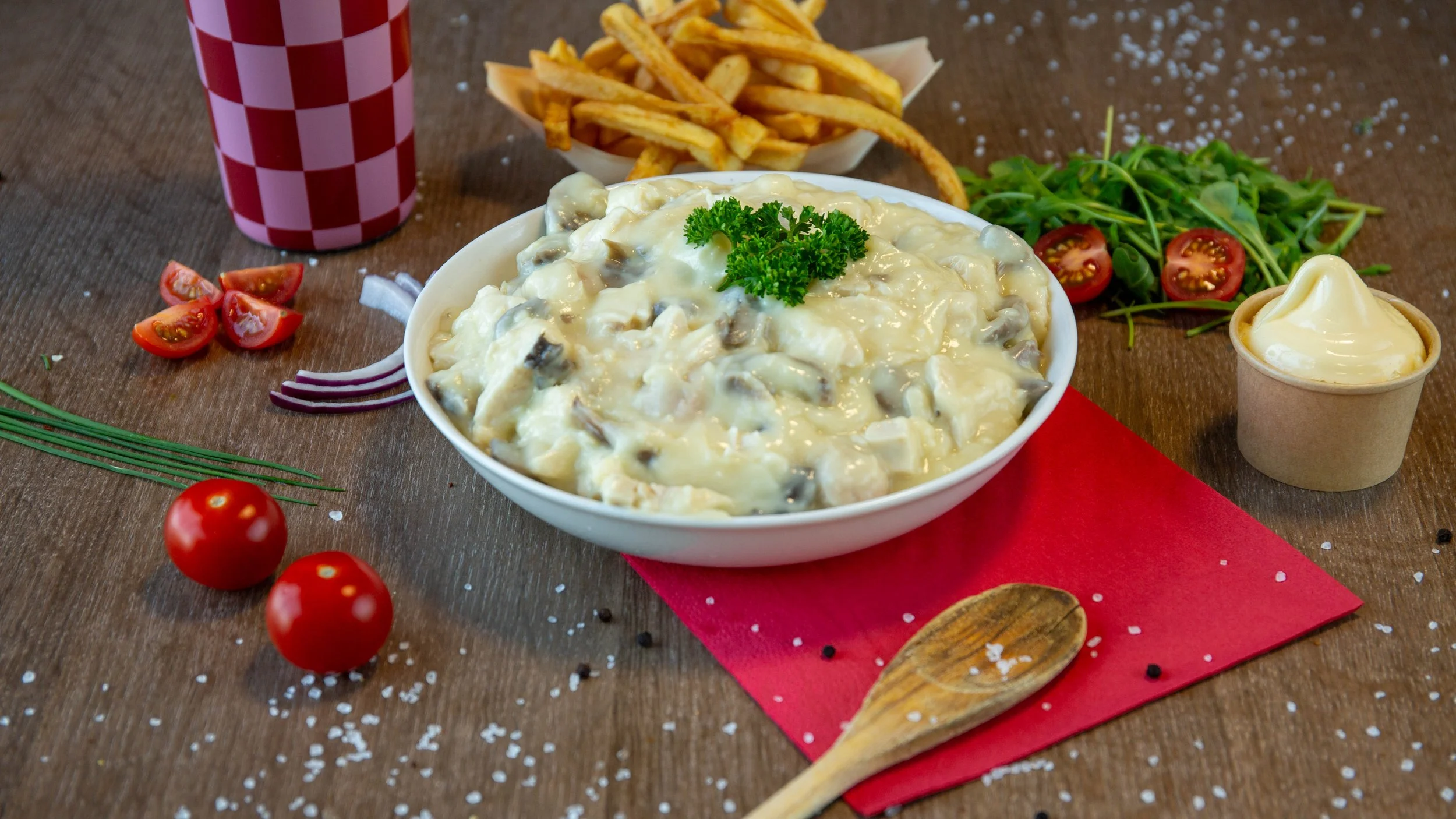Bord met aardappelsalade met champignons, een beker friet, cherrytomaten, sla met tomaatjes, een bekertje mayonaise, en groenten, op een houten tafel.
