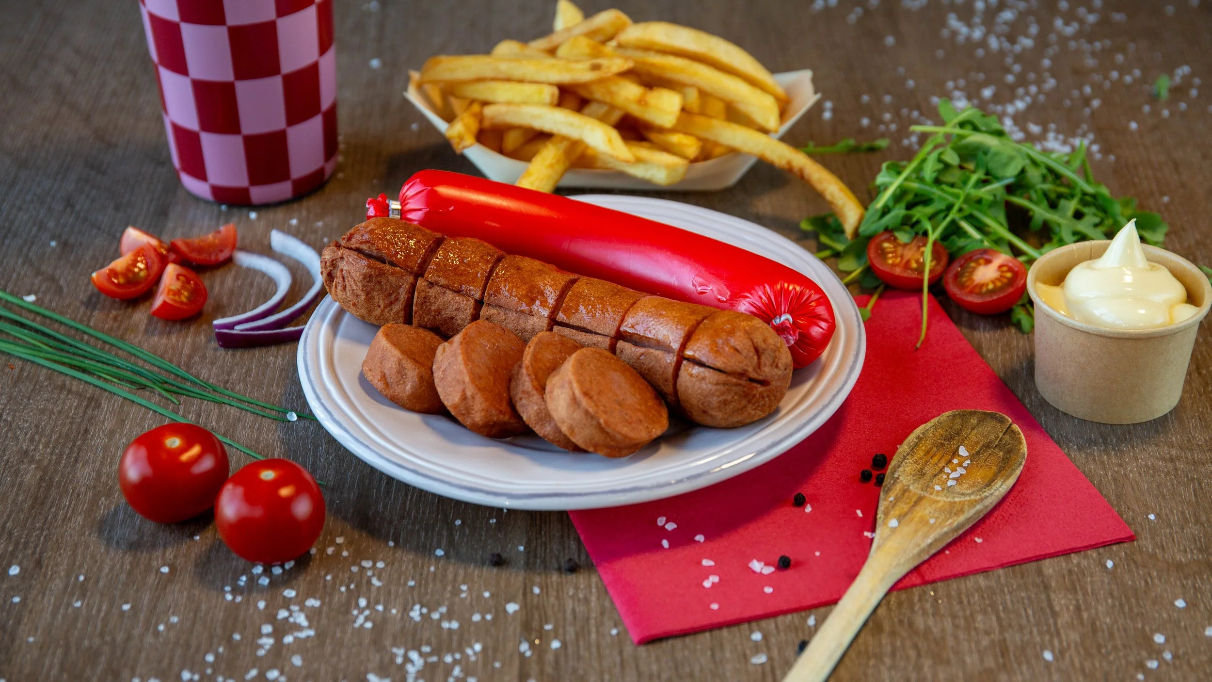 Verschillende gerechten op een houten tafel met tomaten, ijsbergsla, gekookte worst, frietjes, saus, een rode paprika, en een glas met frisdrank.
