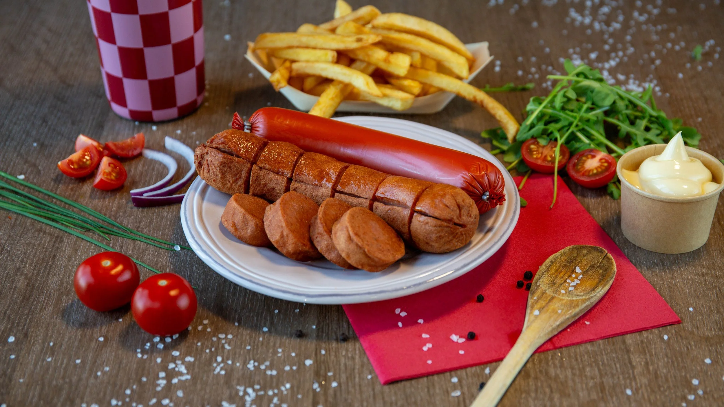 Nederlandse maaltijd met rookworst, gehaktbal, friet, salade, tomaten, rode ui, kerriesaus, en een frisdrank op een houten tafel.