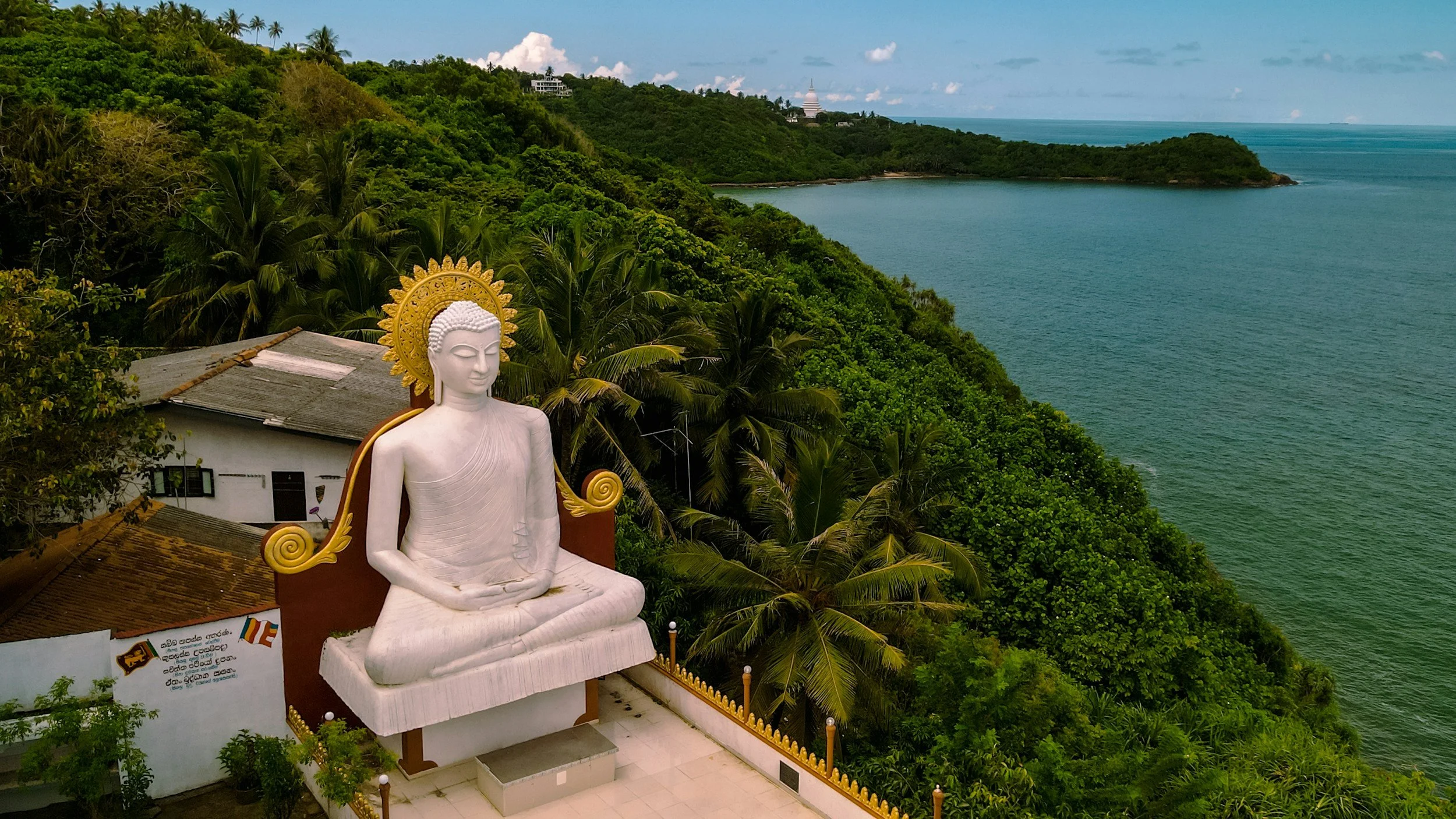 A large white Buddha statue with a golden halo, seated in a meditative pose, overlooking lush green hills and the ocean.